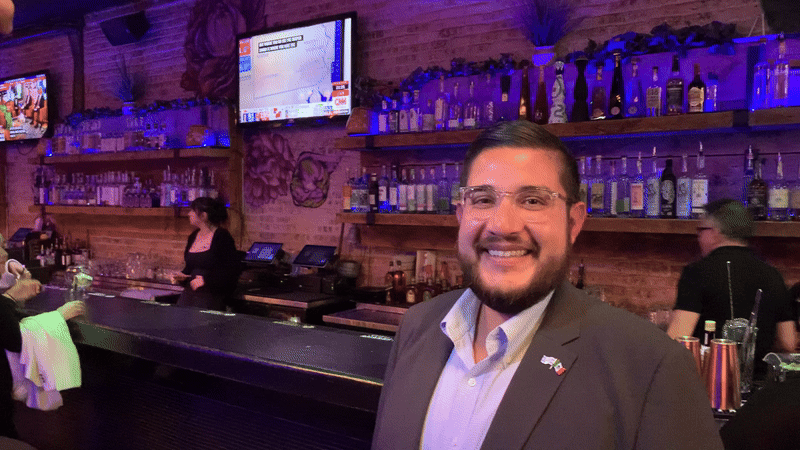 Man in blazer and white button down smiling in front of a bar.