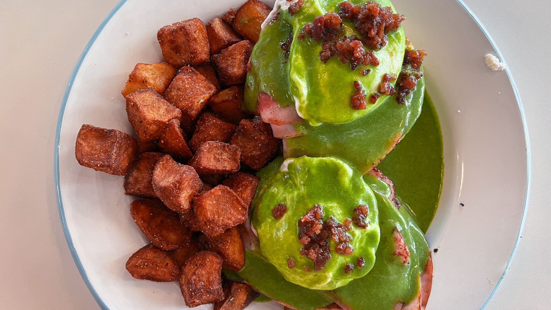 Poached eggs layered on ham and biscuits with a green sauce, served on a plate with cubed potatoes.