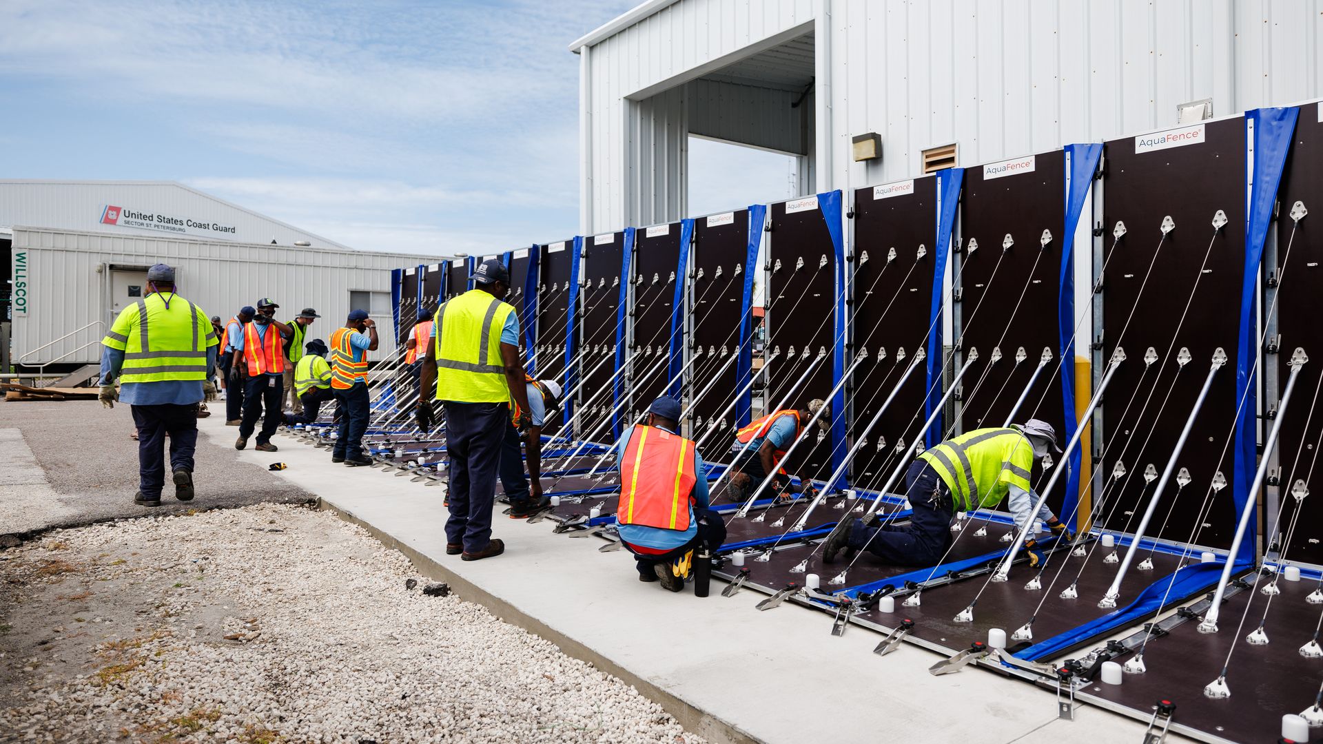 Workers in orange and yellow safety vests assemble large blue and black barriers labeled AquaFence outside a United States Coast Guard building under a partly cloudy sky.