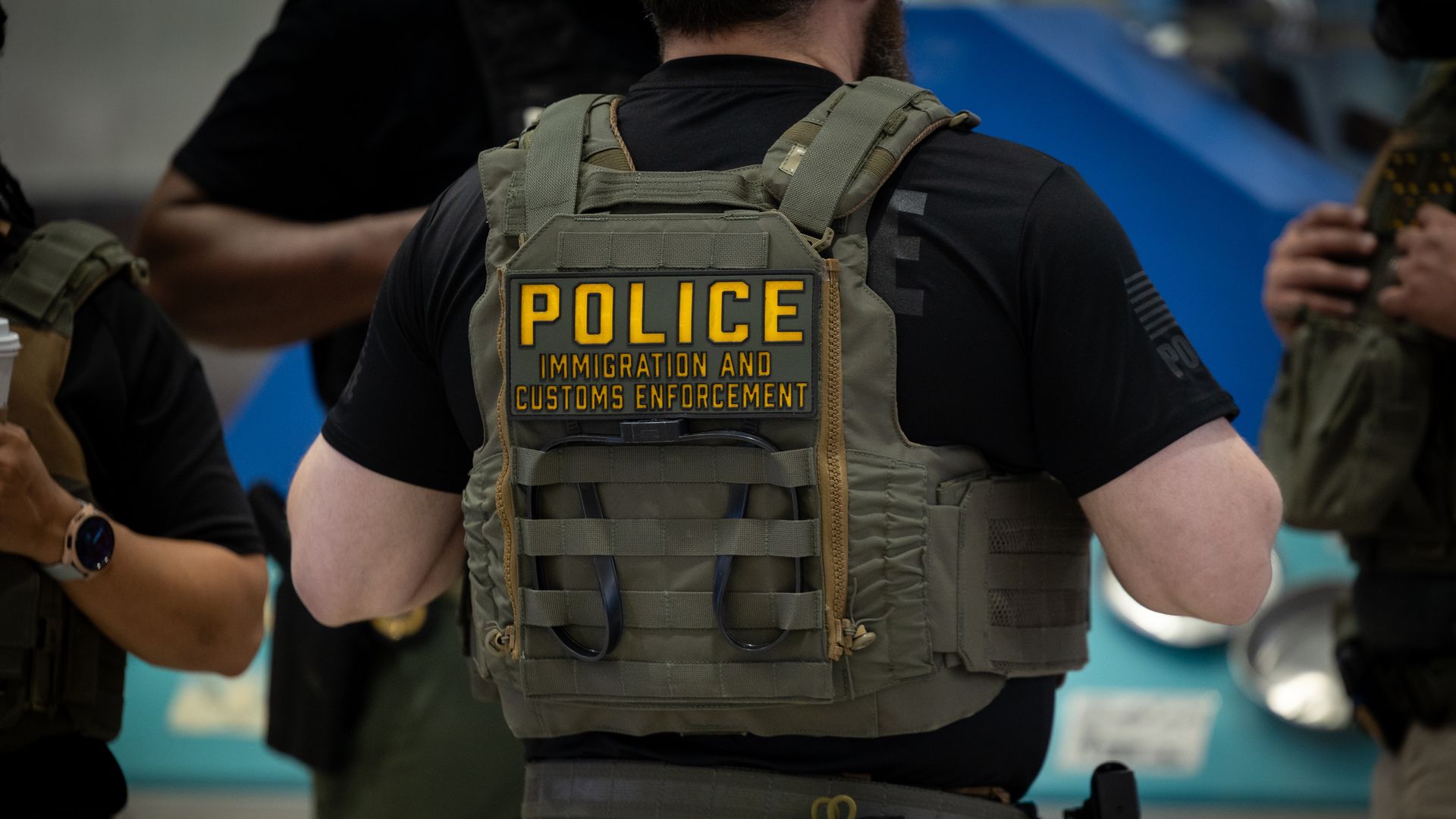 Back view of a police officer wearing a tactical vest with a yellow POLICE patch reading 'IMMIGRATION AND CUSTOMS ENFORCEMENT'; other officers in gear are visible in the background.