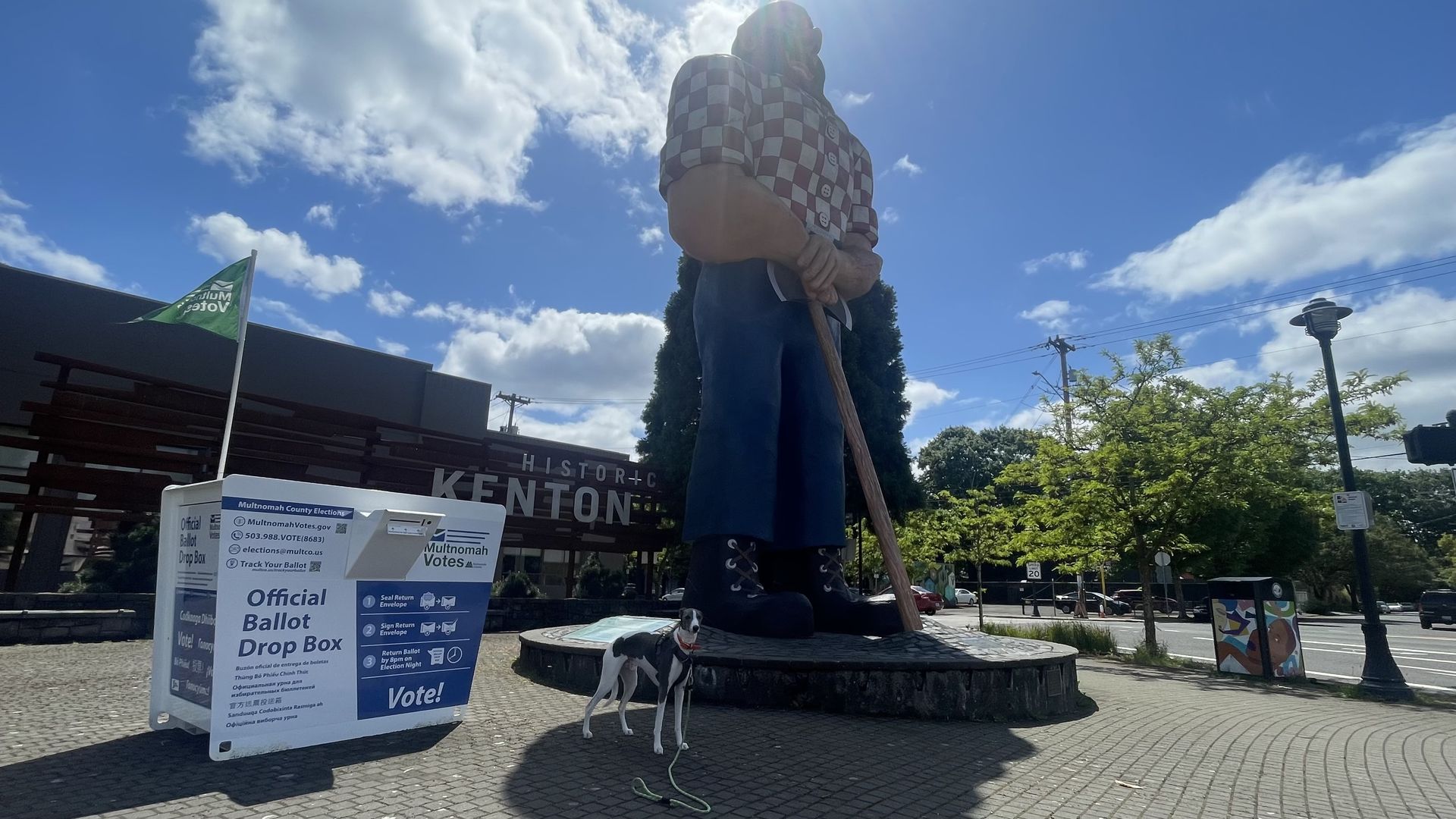 A photo of a dog standing underneath a giant statue of Paul Bunyan and beside a ballot drop box on a sunny day.