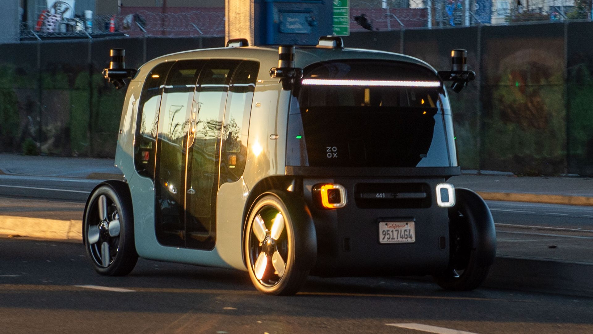 Zoox autonomous taxi with a white, boxy rounded body, large glass doors, four oversized wheels, rooftop sensors and a front LED bar, driving on a city street at sunset.