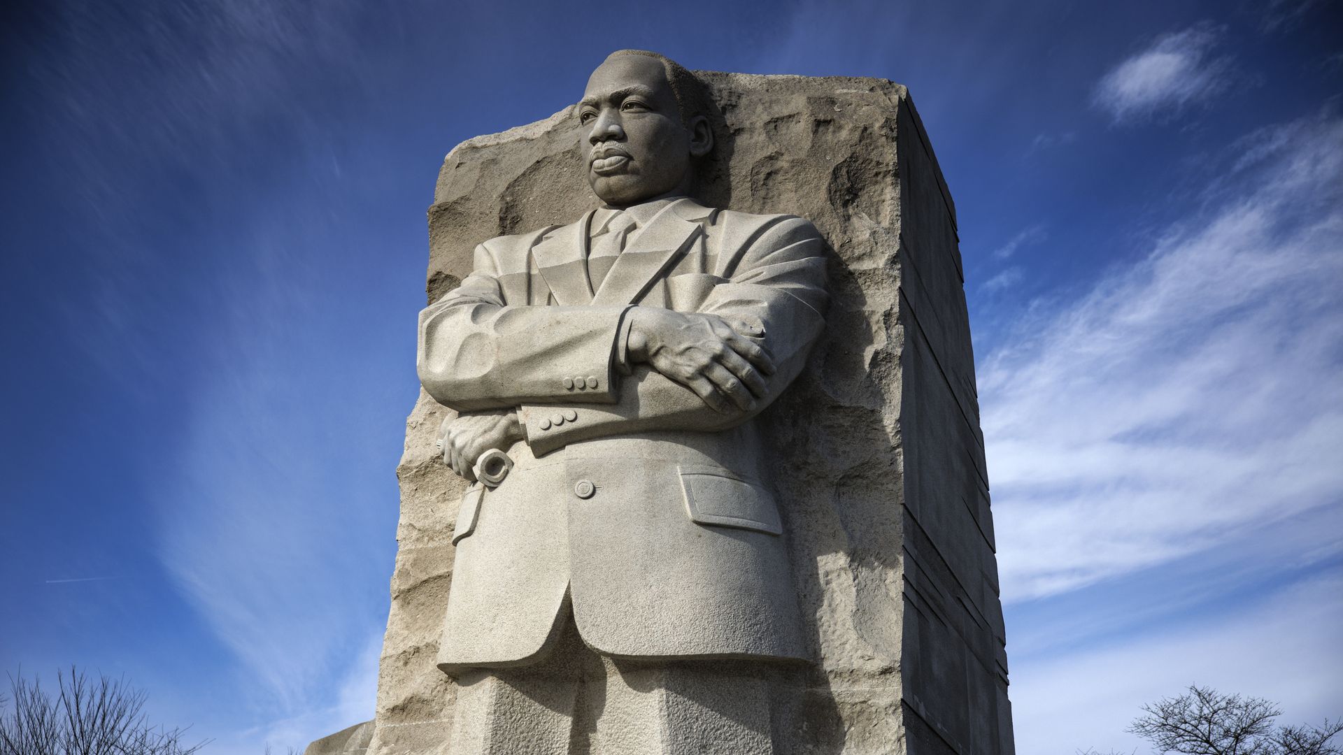 Stone statue of Martin Luther King Jr. with arms crossed, carved from a large block, set against a blue sky with wispy clouds and bare tree branches.