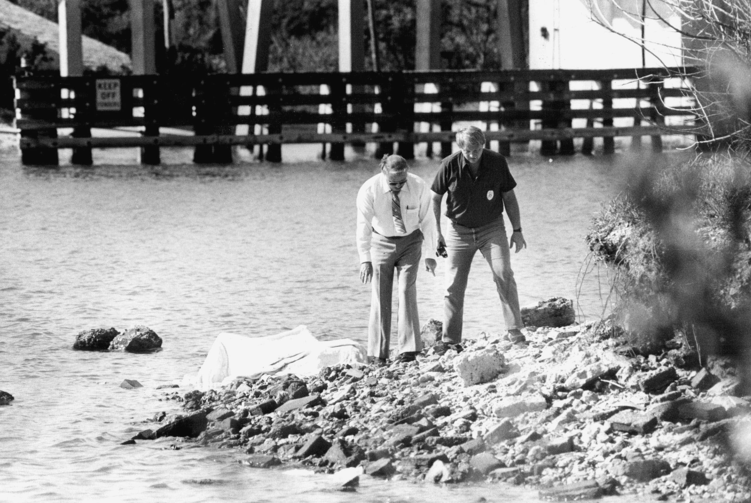 A black-and-white photo showing men standing on a rocky shoreline by water; one wears a white shirt and tie, the other a dark polo with a badge, bending to inspect a white sheet on the rocks, with a wooden pier in the background.