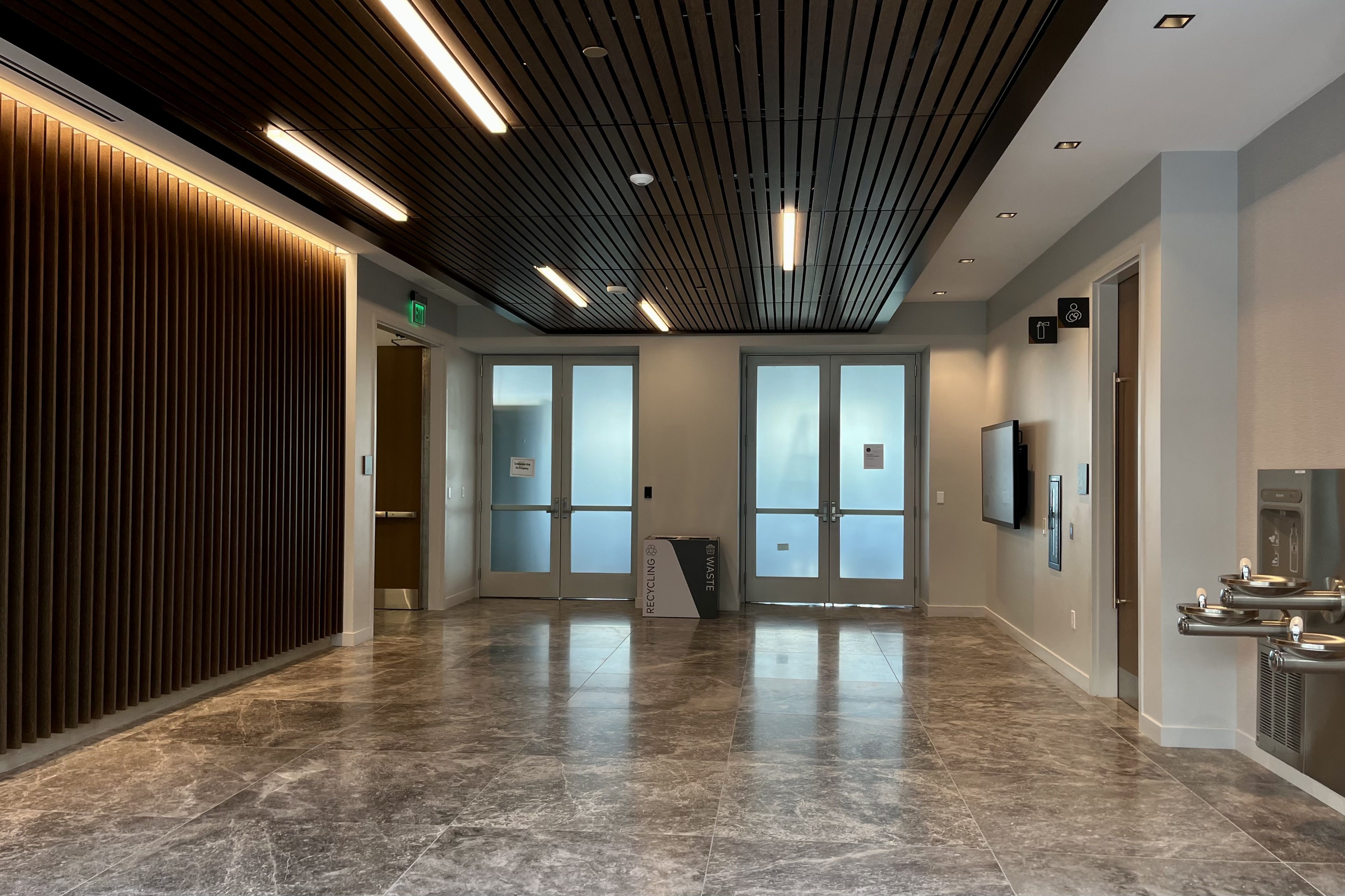 Modern indoor lobby with gray marble floor, wood-paneled ceiling and wall, frosted glass double doors, water fountains on right, and recycling bin between doors.
