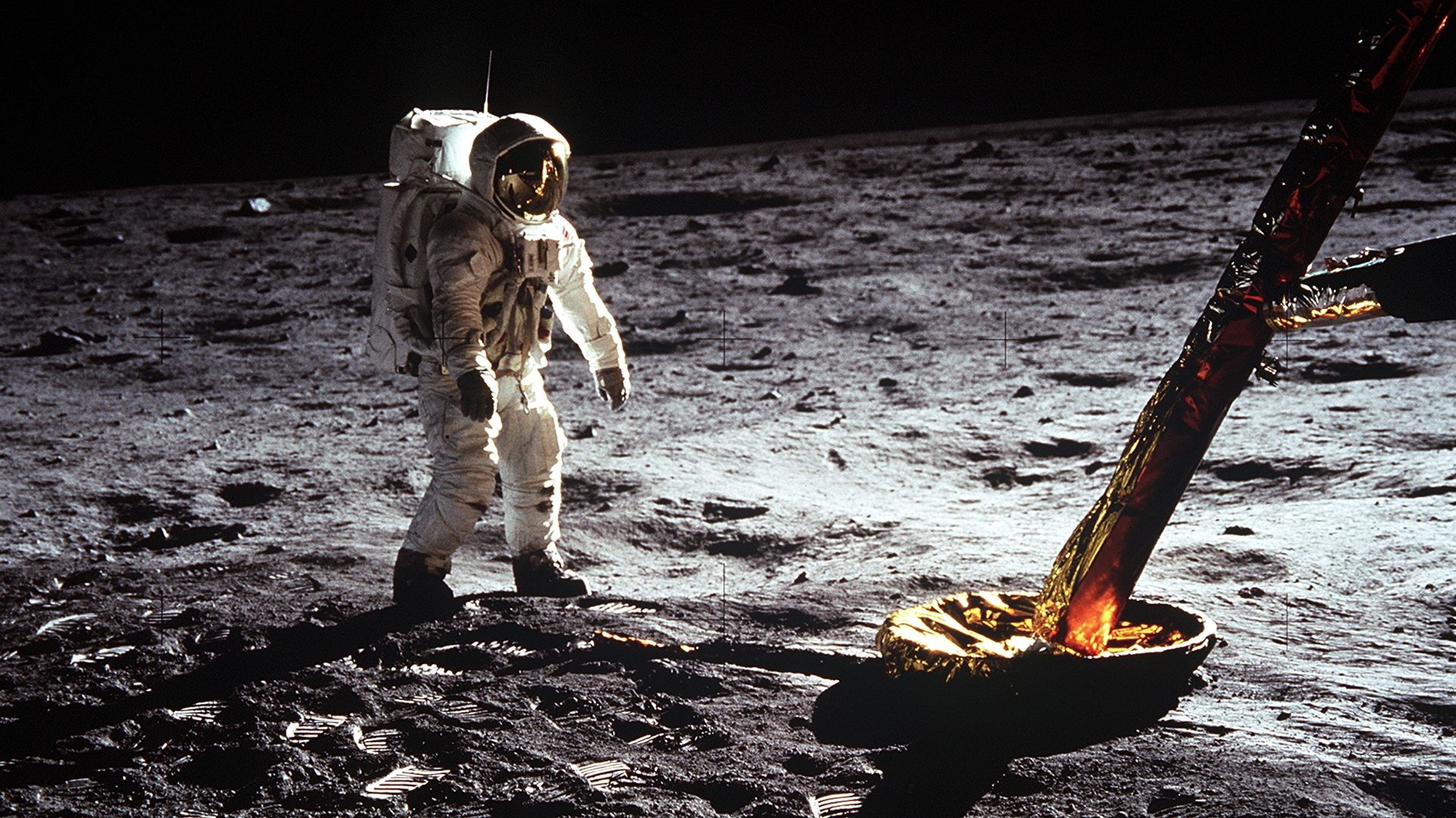 Buzz Aldrin in his EV suit stands next to the leg of the lunar lander on the cratered surface of the Moon