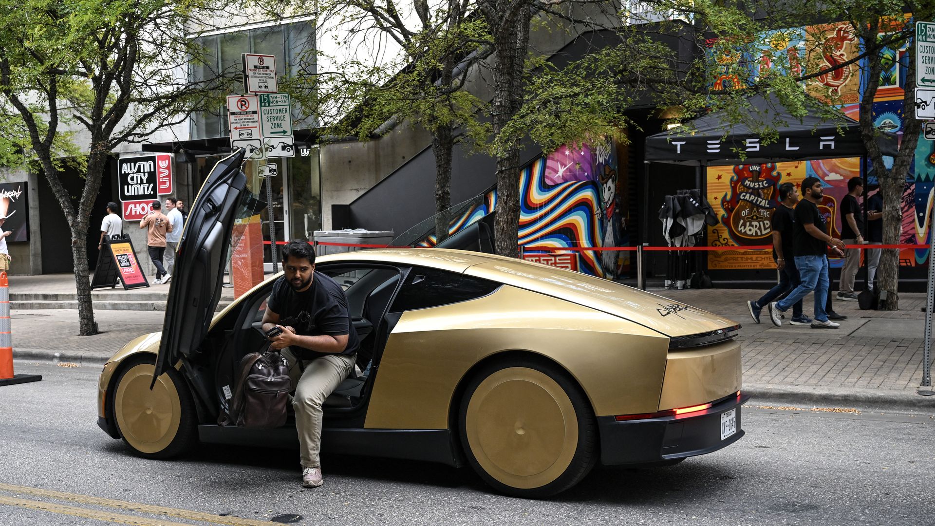 Urban street scene with a gold Tesla car doors open, two men nearby, colorful mural walls, green trees overhead, and city signs and storefronts in the background.