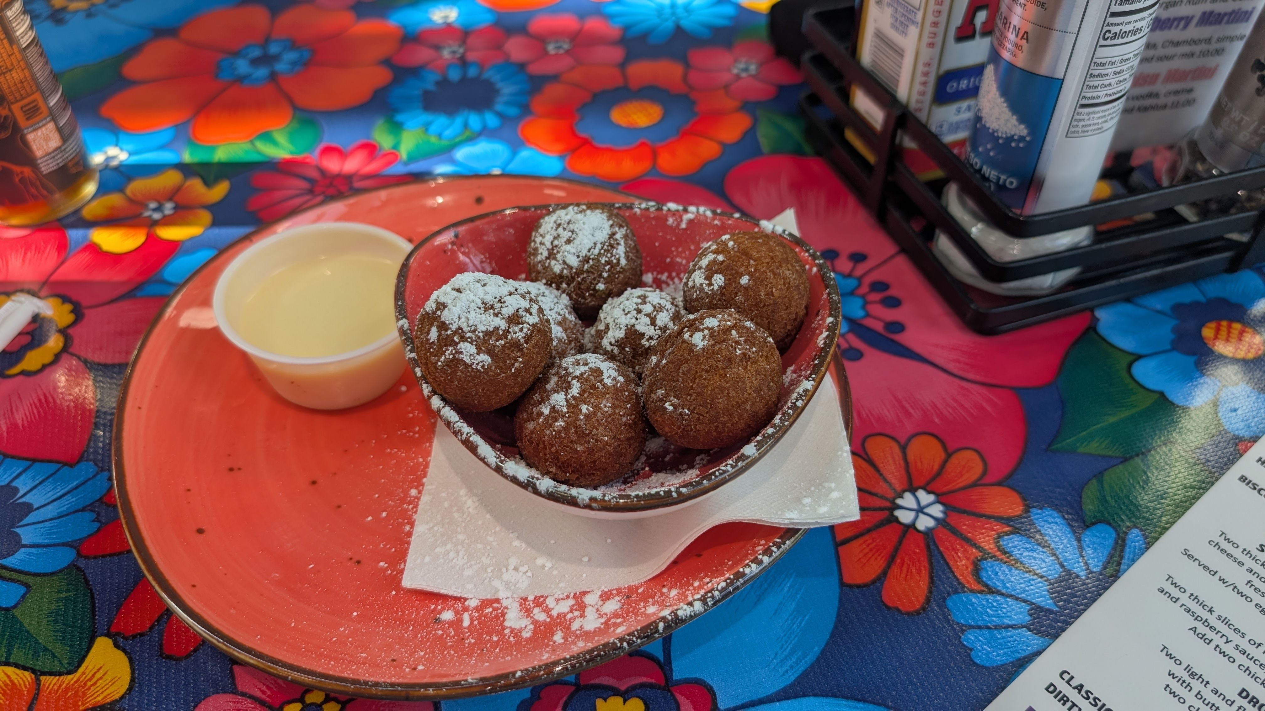 Red bowl with powdered sugar-dusted fried dough balls on a red plate, beside a small cup. Bright blue floral tablecloth with red, orange, and blue flowers; beverage cartons visible in background.