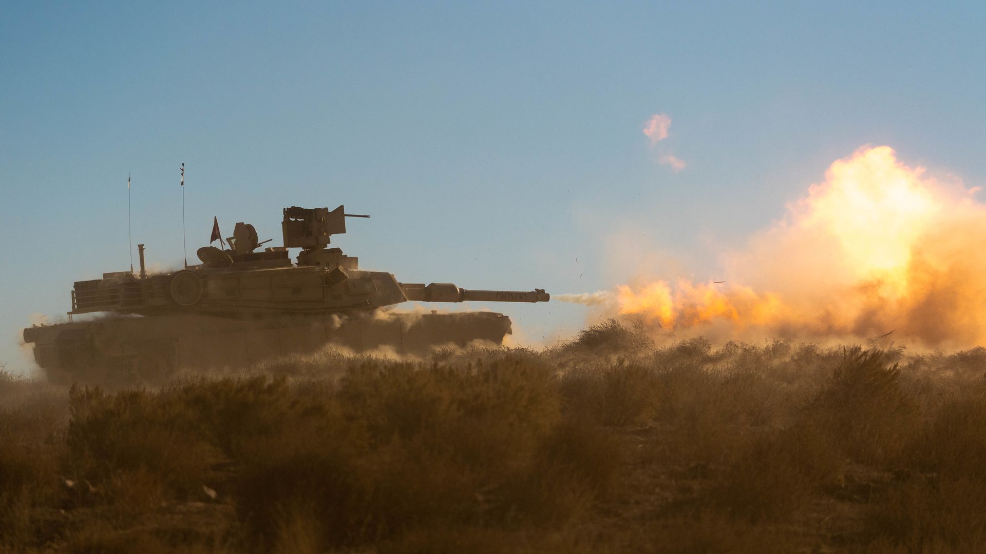 An Abrams tank, at left, fires downrange. A large fireball and dust cloud can be seen, at right. The sky above is blue.
