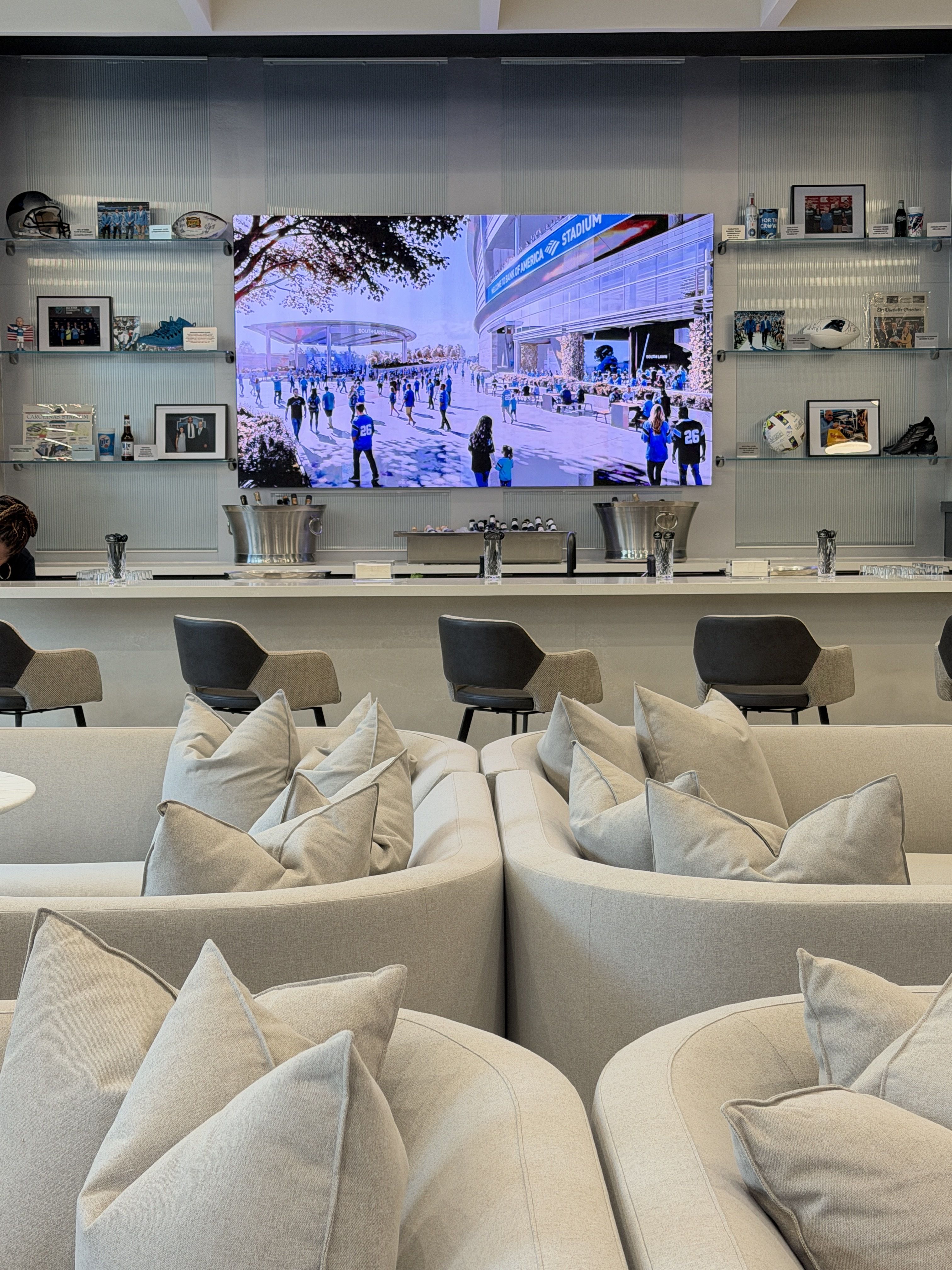 Modern lounge with pale beige curved sofas and cushions, facing a large wall-mounted screen showing a bright outdoor stadium scene with people; glass shelves flank wall with photos and memorabilia.