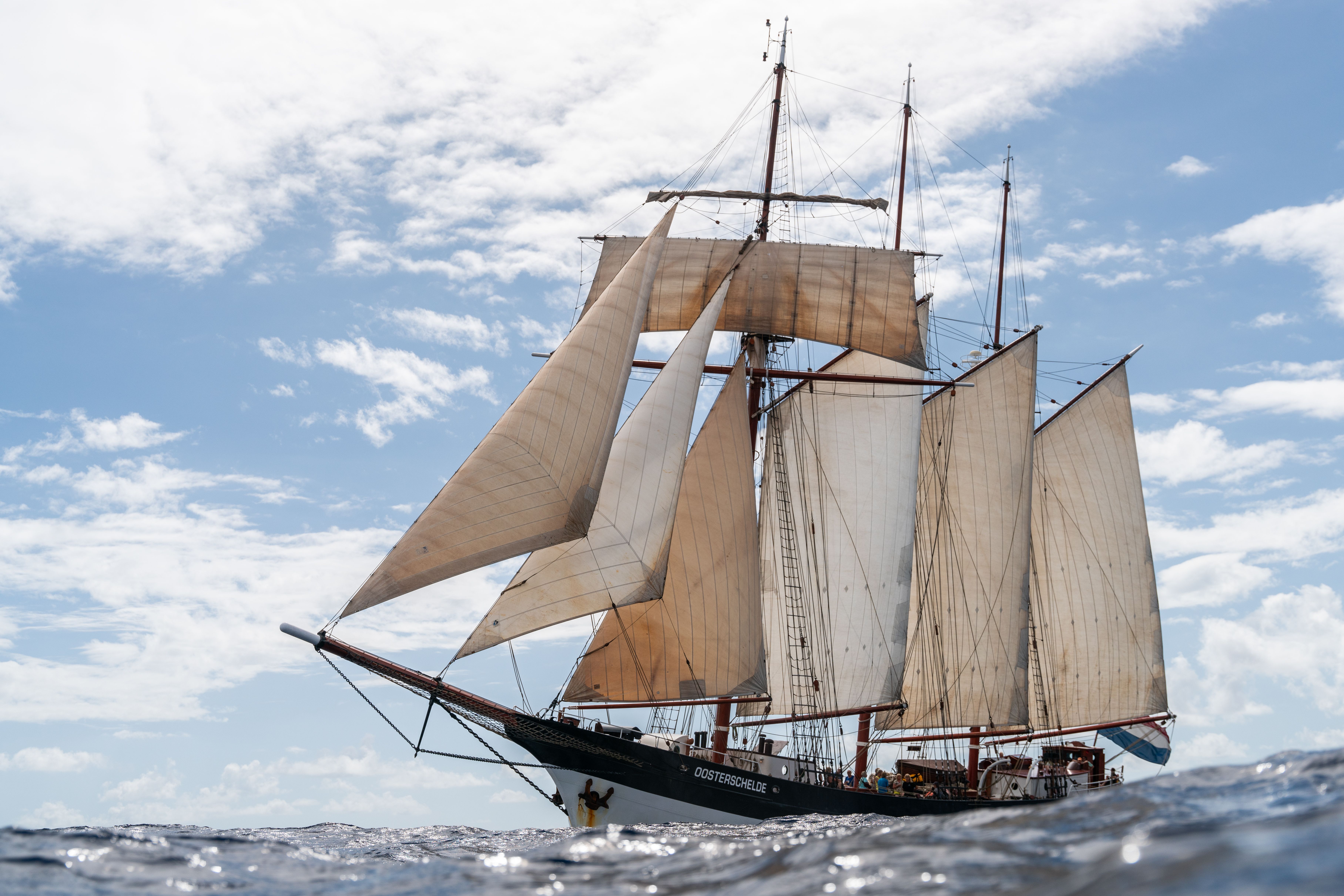 Tall sailing ship with cream canvas sails, brown masts and complex rigging, gliding on a blue ocean under a partly cloudy sky; crew visible on the deck.