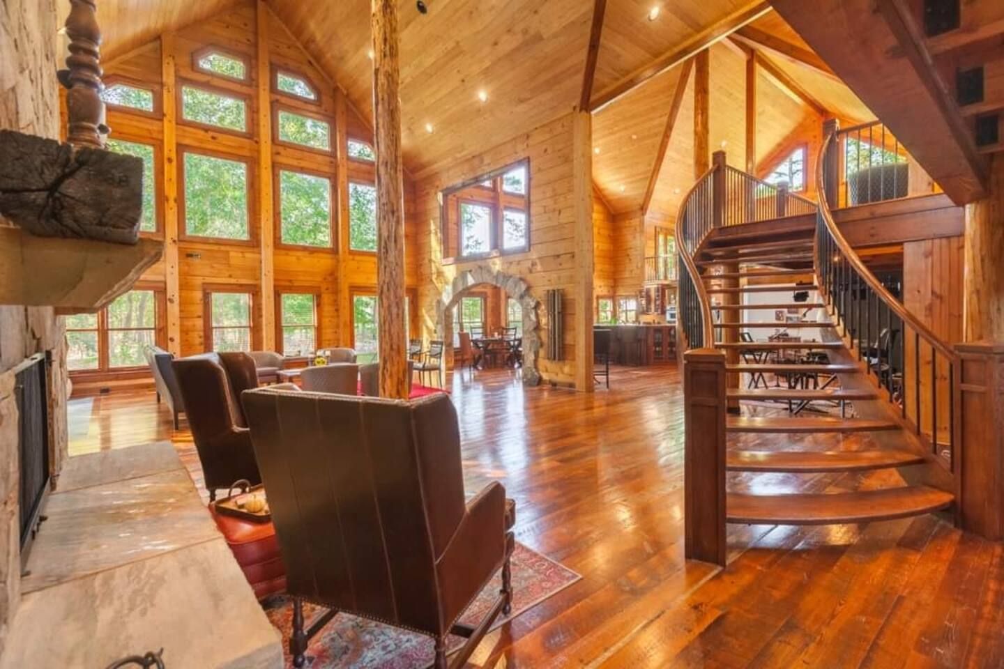 Spacious rustic living area of a wooden cabin with high ceilings, large windows, stone fireplace, leather chairs, curved wooden staircase, and warm hardwood floors with natural light.