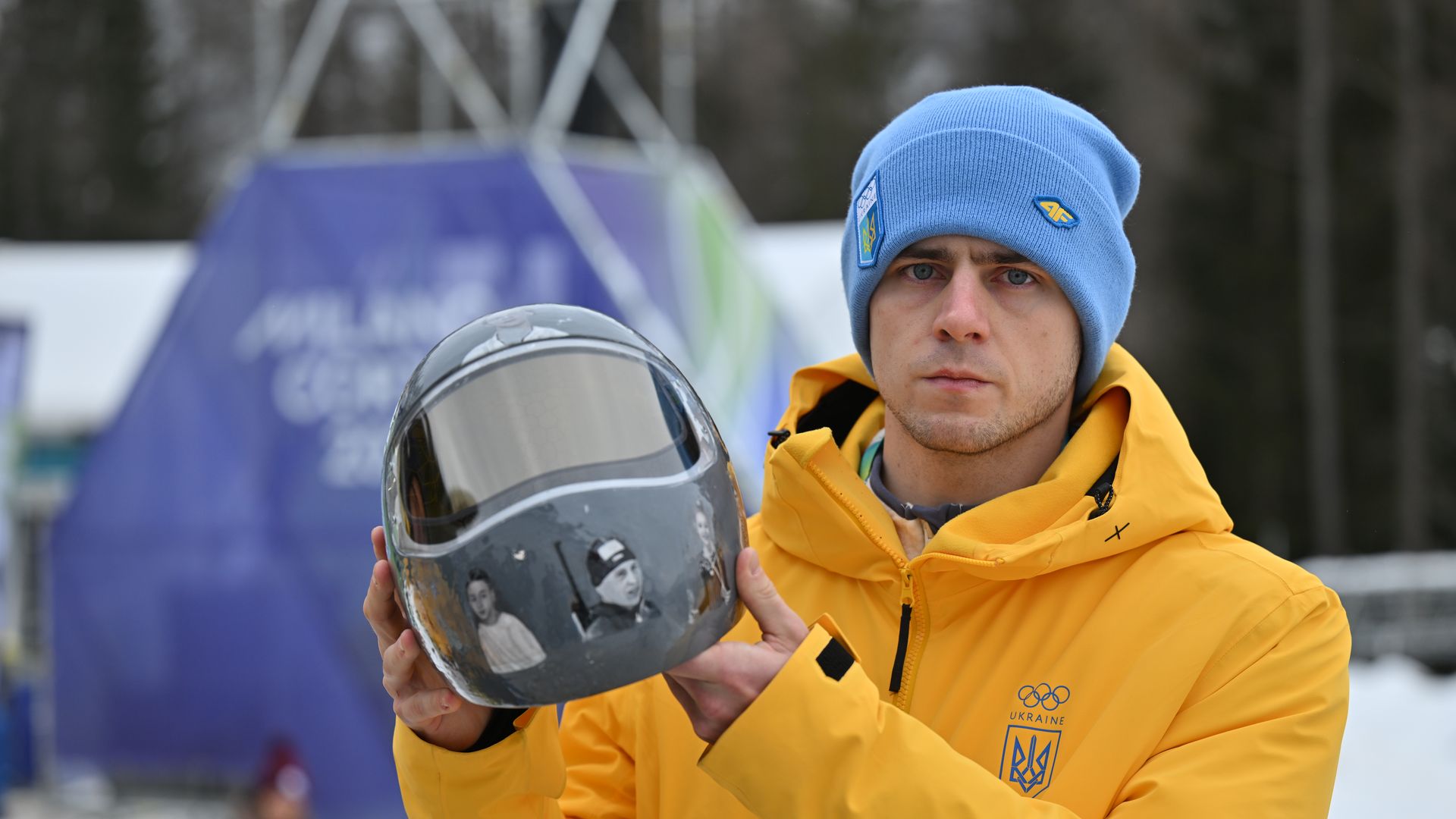 Heraskevych standing outside wearing a bright yellow jacket and blue beanie while holding his helmet which has photos of athletes and coaches killed in the Russia-Ukraine war.