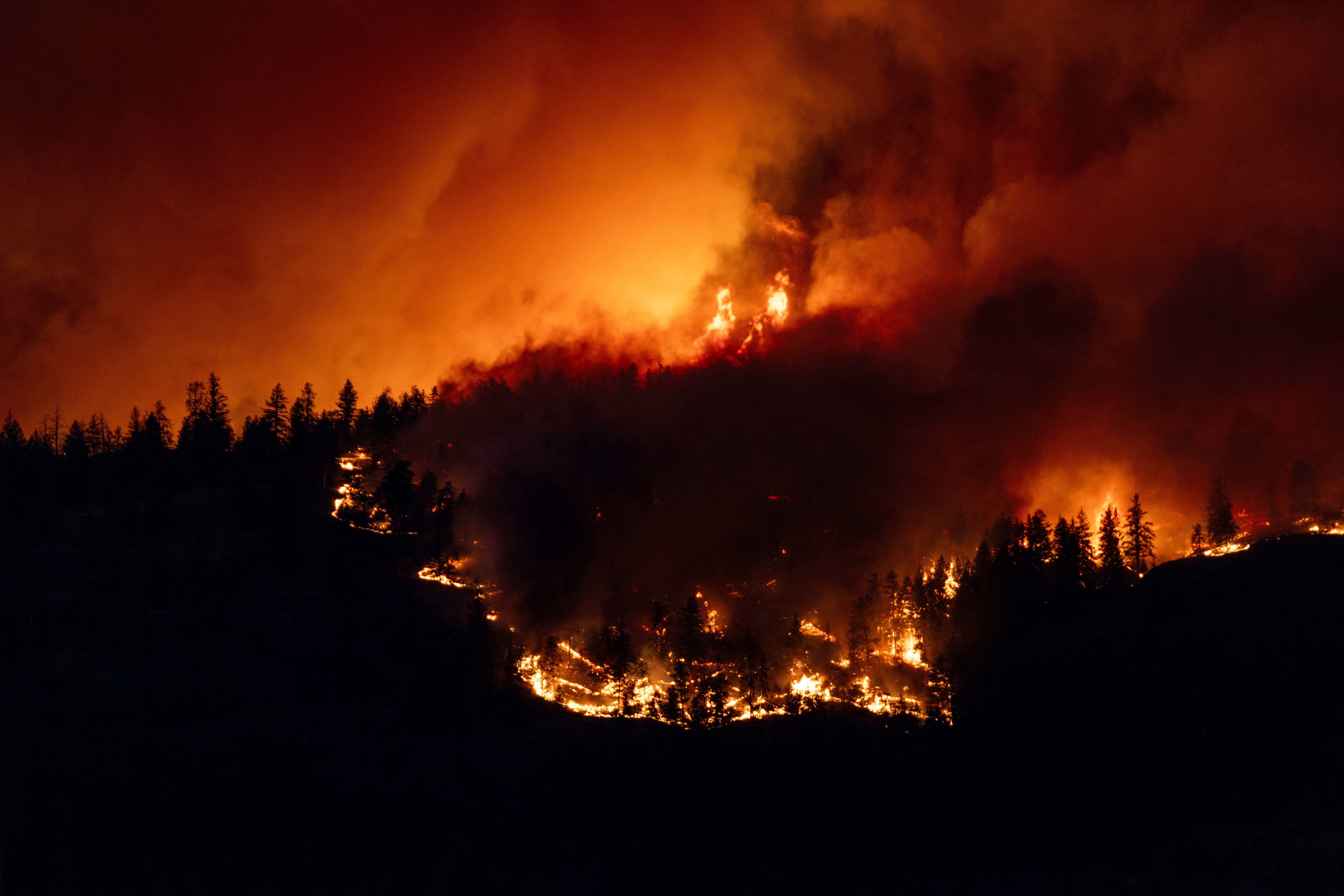 A night time photo showing a fire burning along a mountain ridge. Nothing is visible except for the outlines of trees against the fire and smoke. 