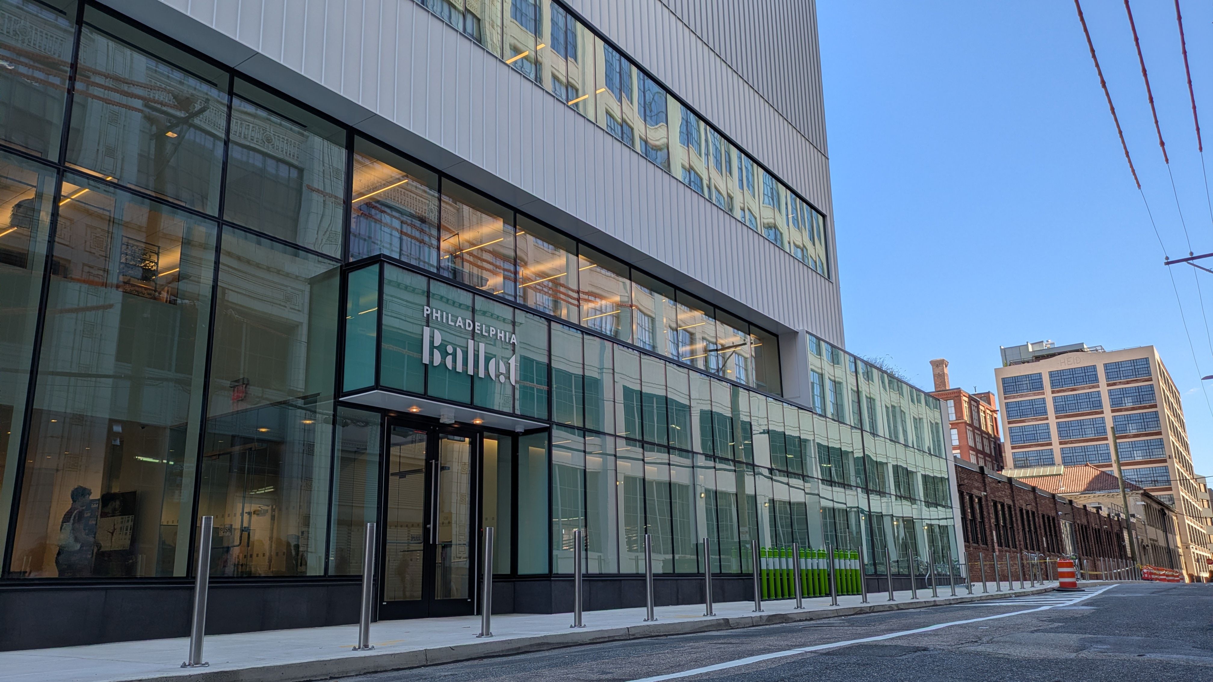 Modern glass-front building with a large sign reading "Philadelphia Ballet" on the lower level. Reflections of a historic facade in the glass, blue sky, and a street with bollards and an orange construction barrel.