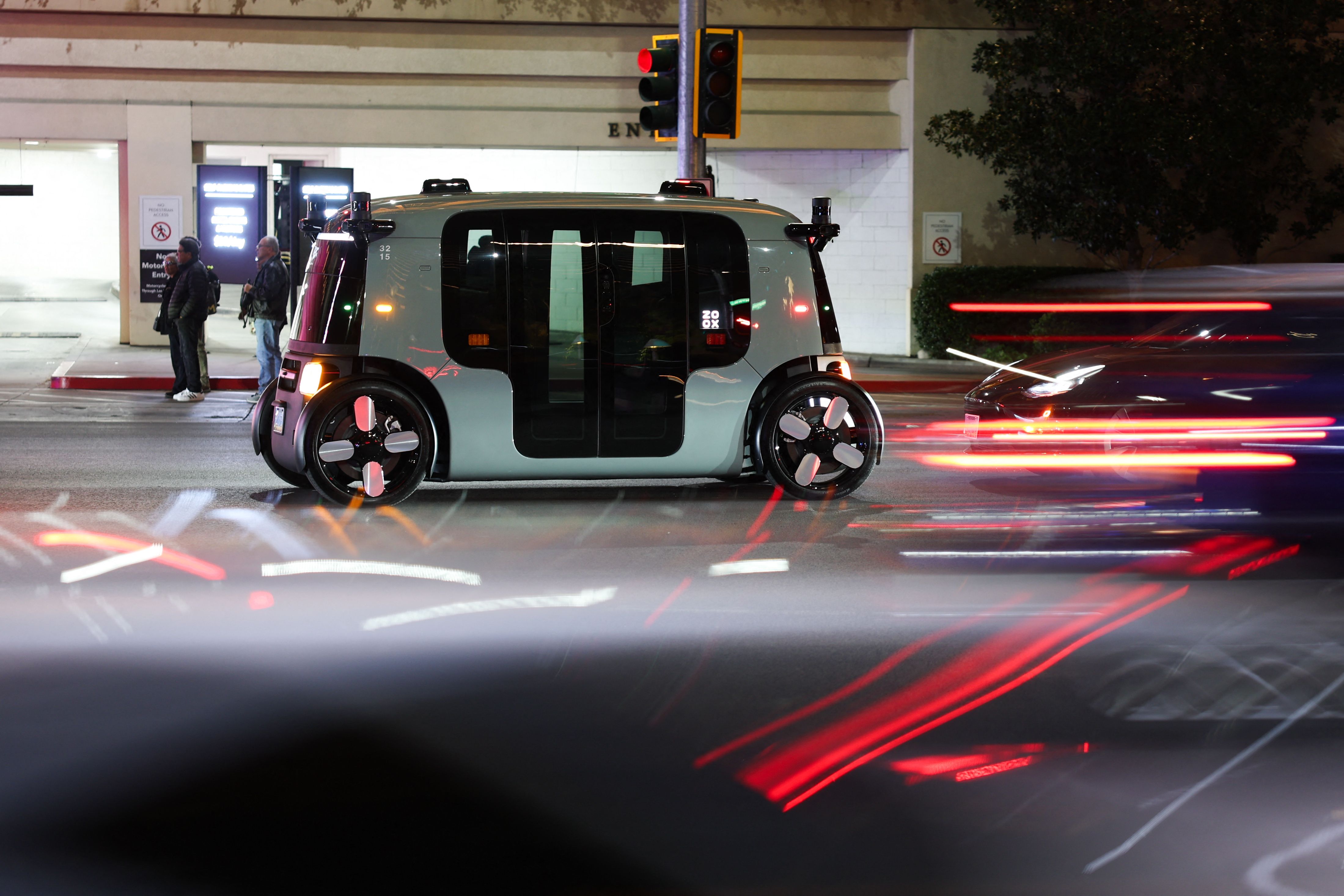 A pale blue-gray autonomous shuttle with oversized wheels and dark glass sides sits on a city street at night, while red light-trail car traffic blurs past; two pedestrians stand on the sidewalk.