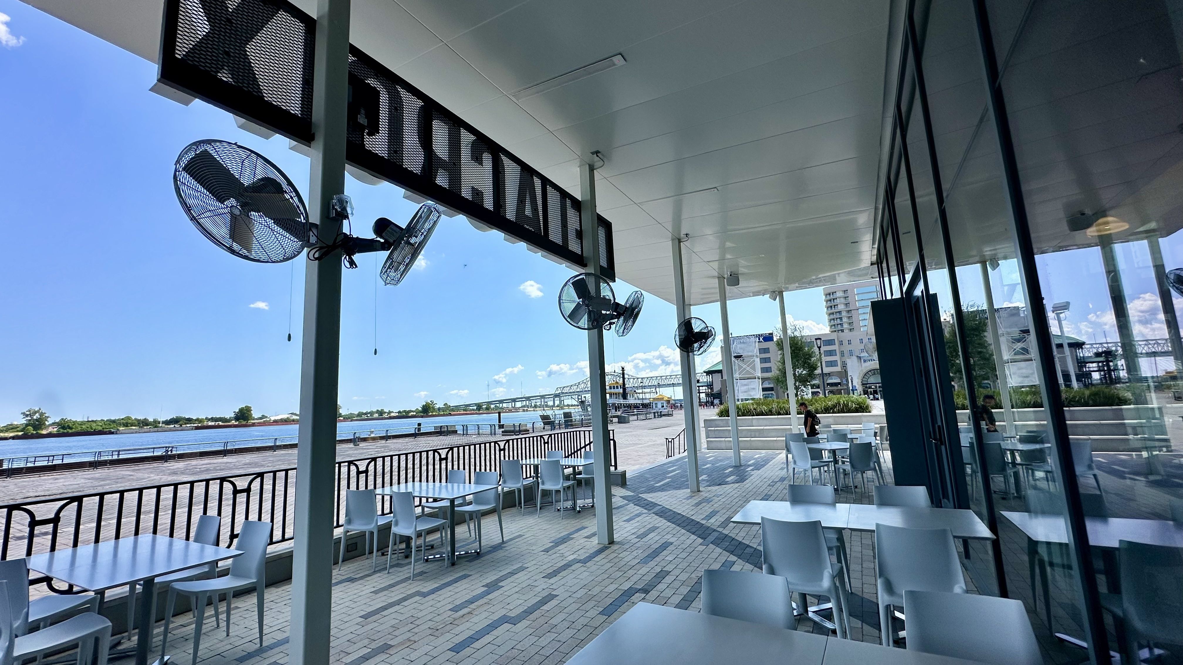 Outdoor seating area with white tables and chairs under a covered patio by the waterfront on a sunny day, with fans on poles and a view of a bridge and buildings across the river.