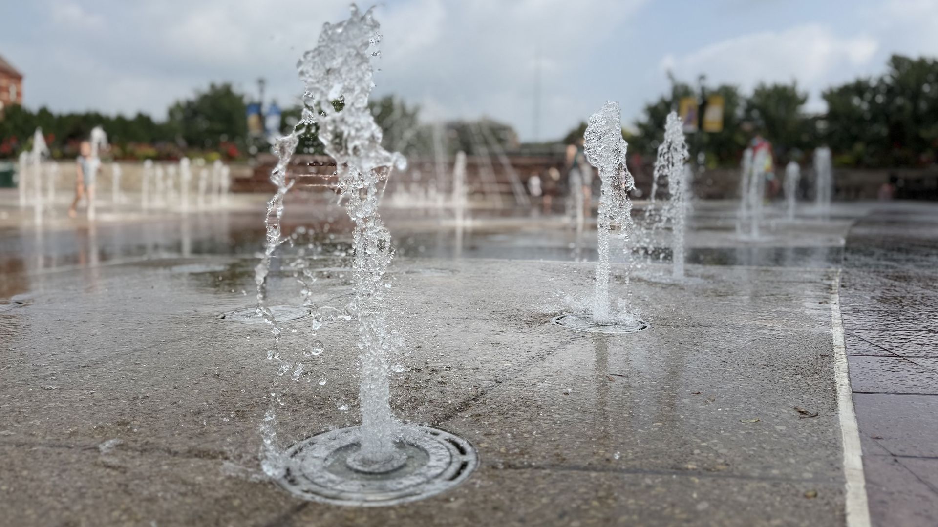 Water jets rise from circular basins in a wet plaza, with blurred people and trees in the background under a blue sky with clouds.