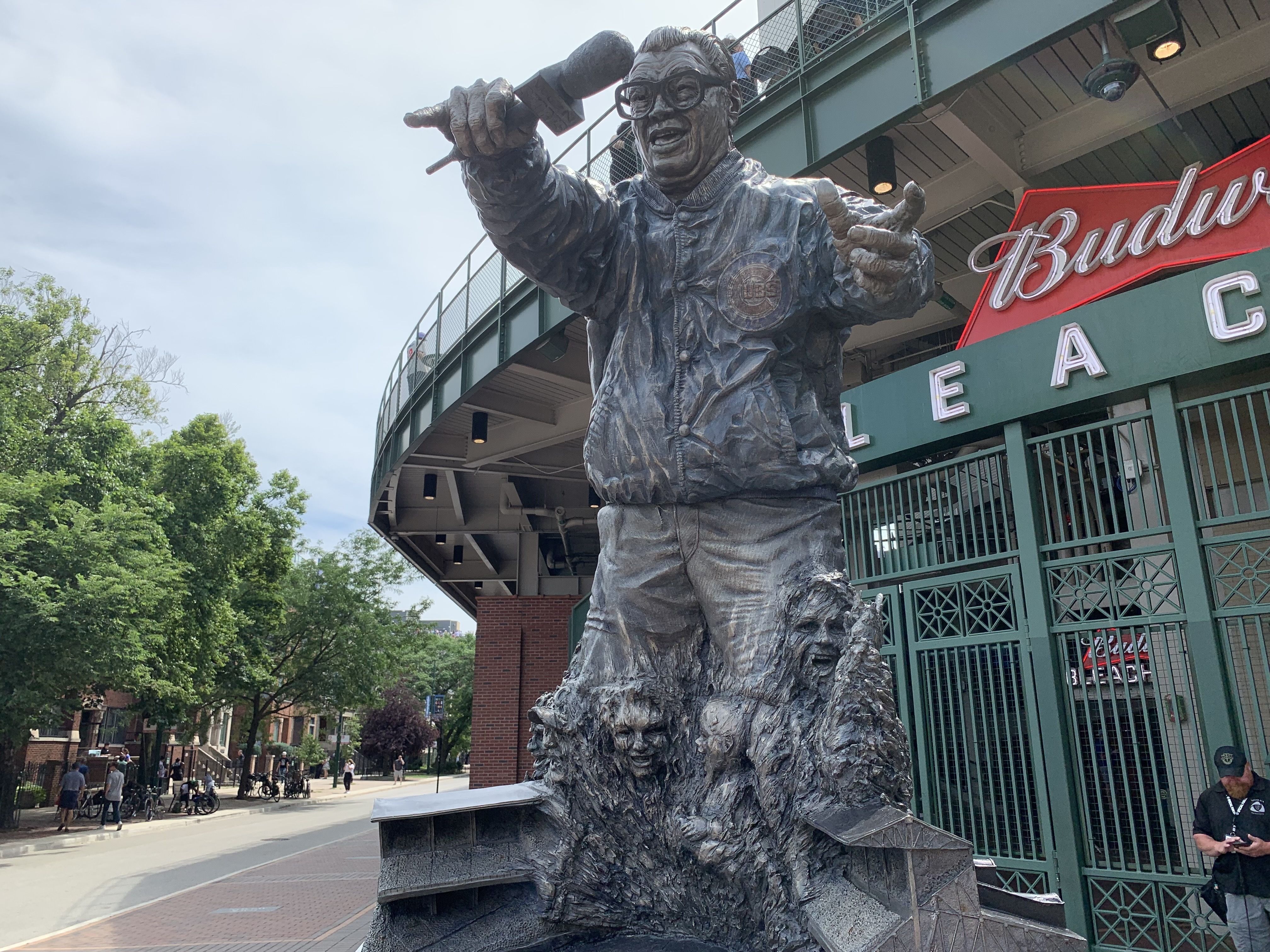 Bronze statue of a man with glasses holding a microphone, surrounded by three faces and miniature stadium seats, in front of a Budweiser sign at a ballpark entrance.