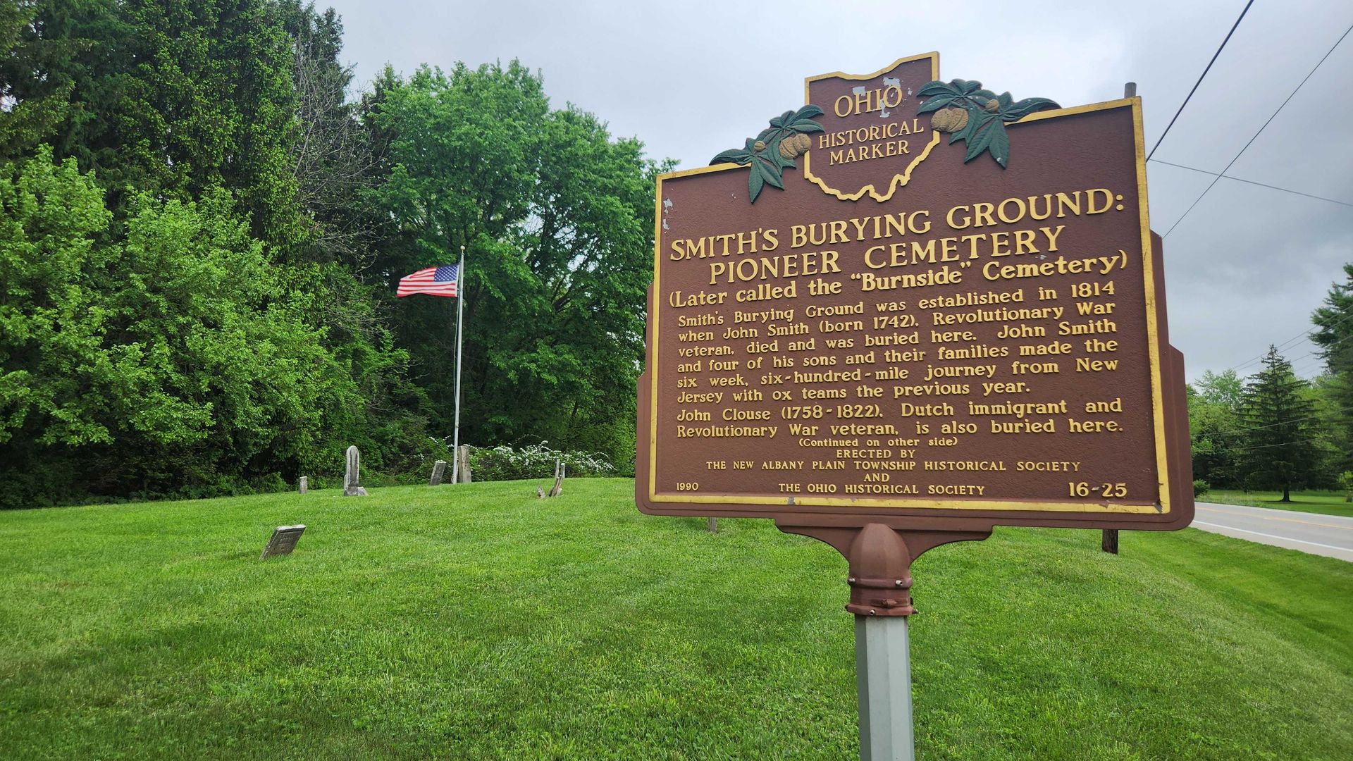 A historical marker for "Smith's Burying Ground: Pioneer Cemetery" with a flag waving in the background and gravestones present. 