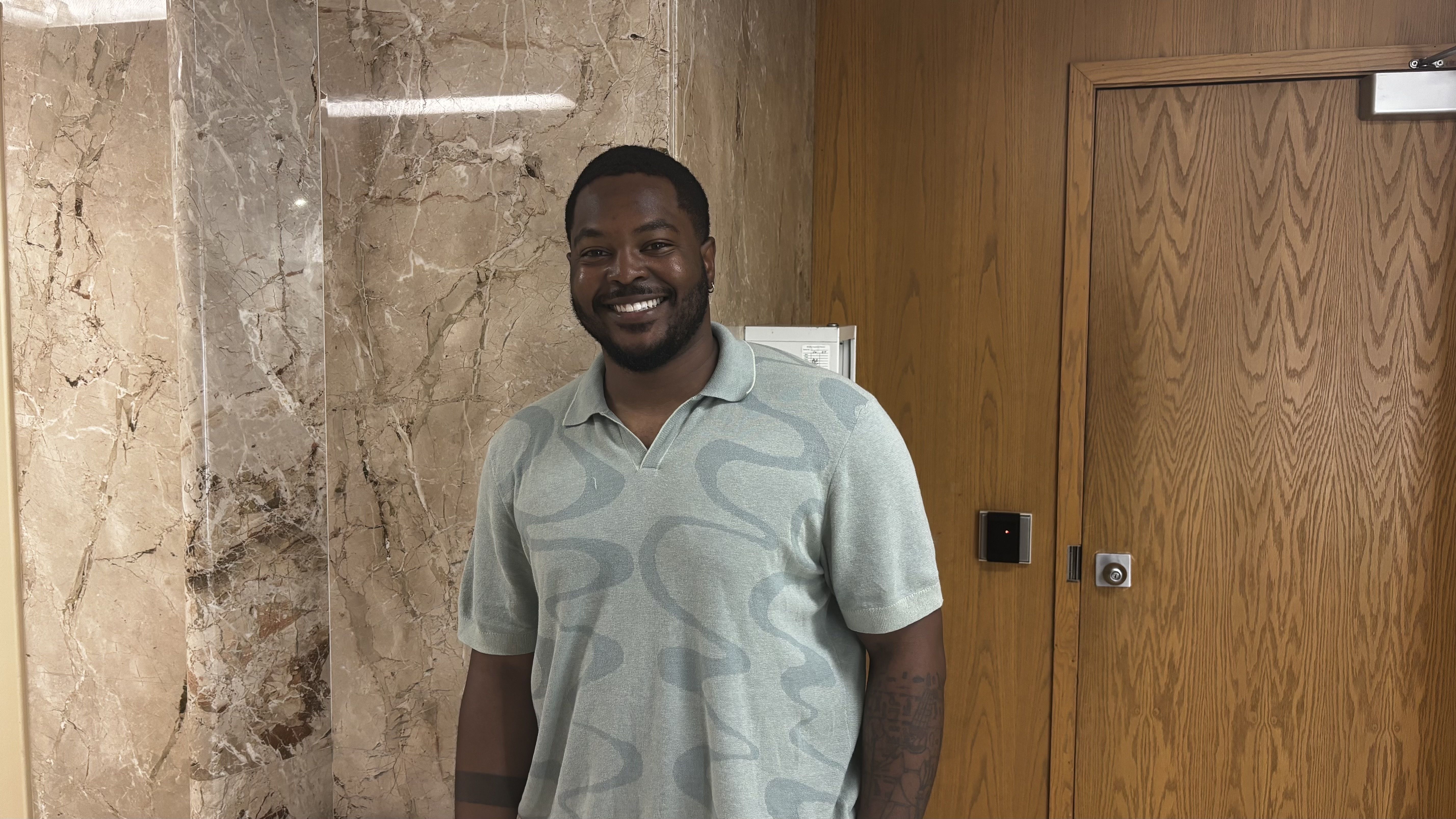 Smiling man with short black hair and a beard, wearing a light green patterned polo, stands in front of a marble wall and a wooden door.