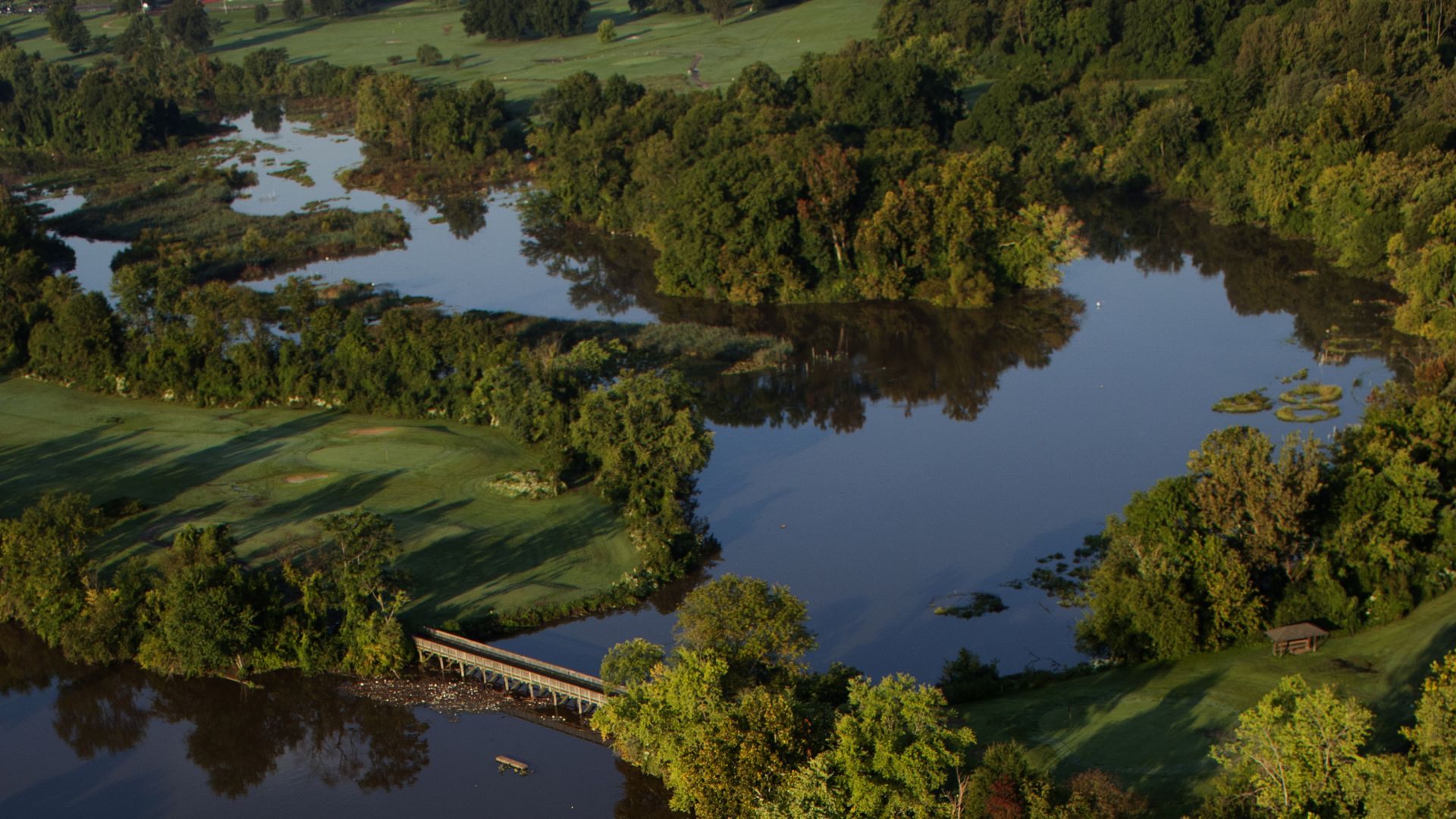 Aerial view of a green golf course bordered by lush trees and two bodies of brown and blue water with a wooden bridge connecting the grassy areas.