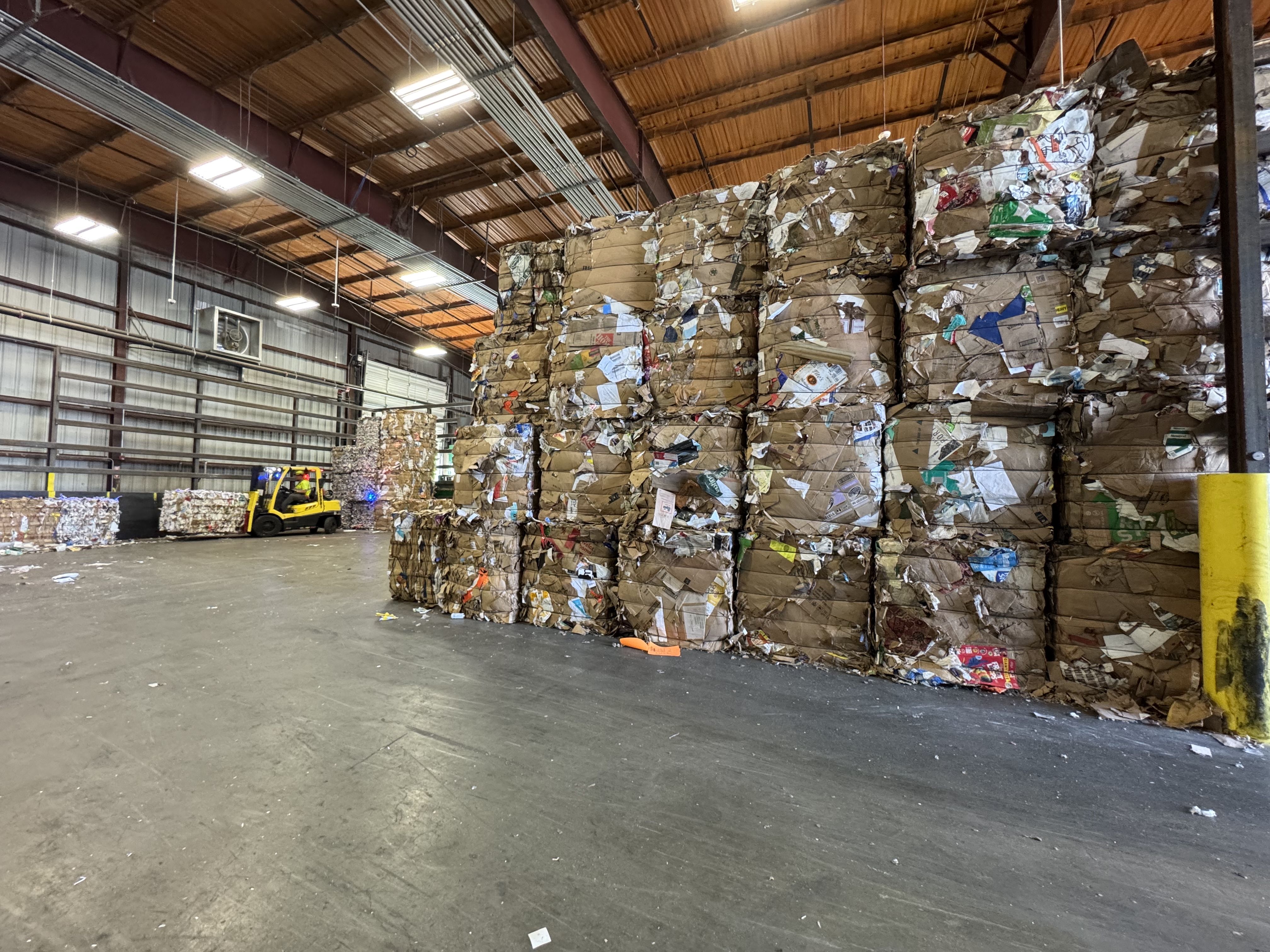 Warehouse with stacked bales of crushed cardboard for recycling and a yellow forklift moving more cardboard in the background under a wooden ceiling with fluorescent lights.