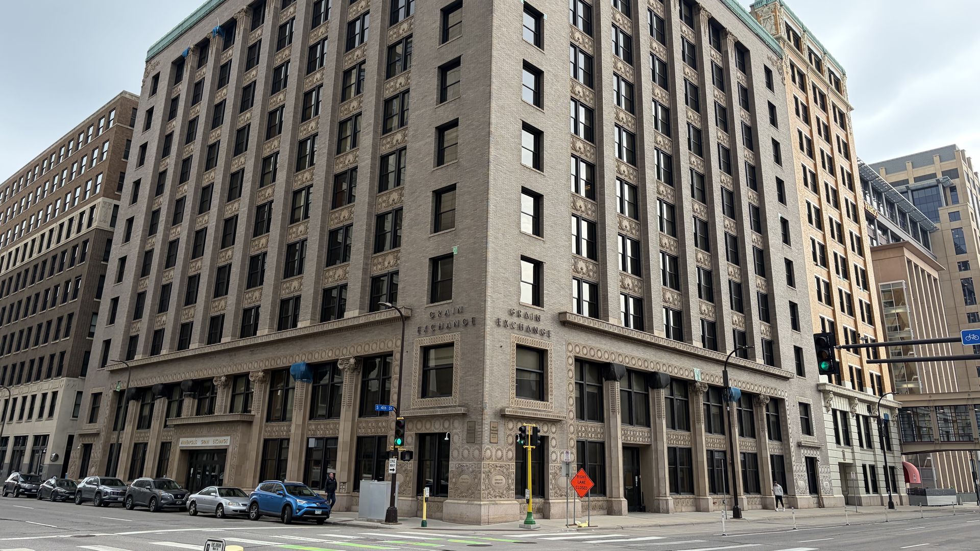 Beige brick corner building with many tall windows and decorative trim at a city intersection; the sign reads "Grain Exchange" as cars line the street and pedestrians pass by.