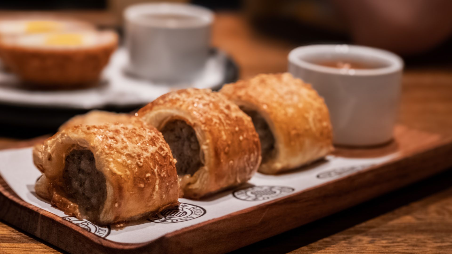 Three golden brown sausage rolls on a wooden board with white paper, accompanied by a small white cup of dipping sauce, set on a wooden table with blurred background items.