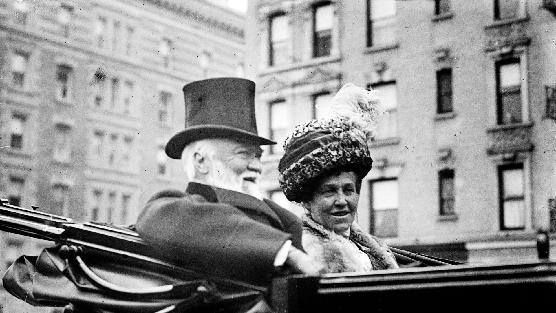 Black-and-white photo of an elderly man in a top hat and coat and a woman in a fur coat with a feathered hat, sitting together in an open carriage on a city street.