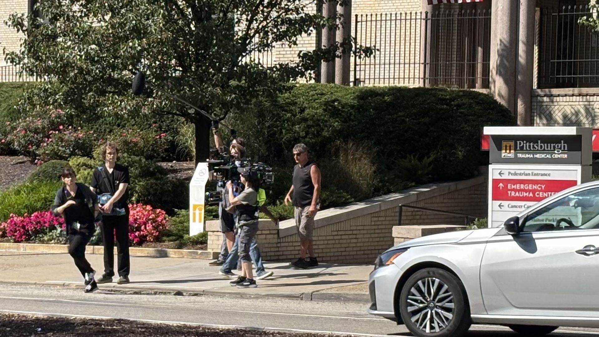 Film crew with camera and boom mic films a person running outside Pittsburgh Trauma Medical Center, surrounded by green bushes, flowers, and a white car parked nearby on a sunny day.