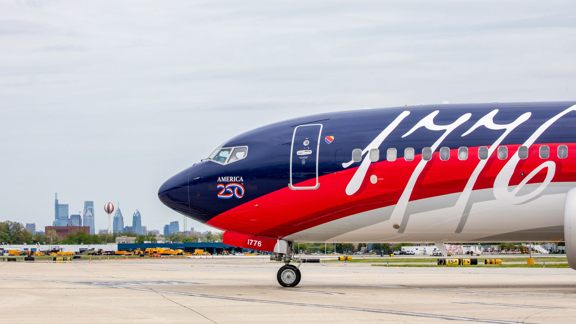 Side view of a large passenger plane on a runway, dark blue nose and red-blue fuselage with white cursive script; "AMERICA 250" badge near cockpit and "1776" on the nose gear, city skyline in distance.