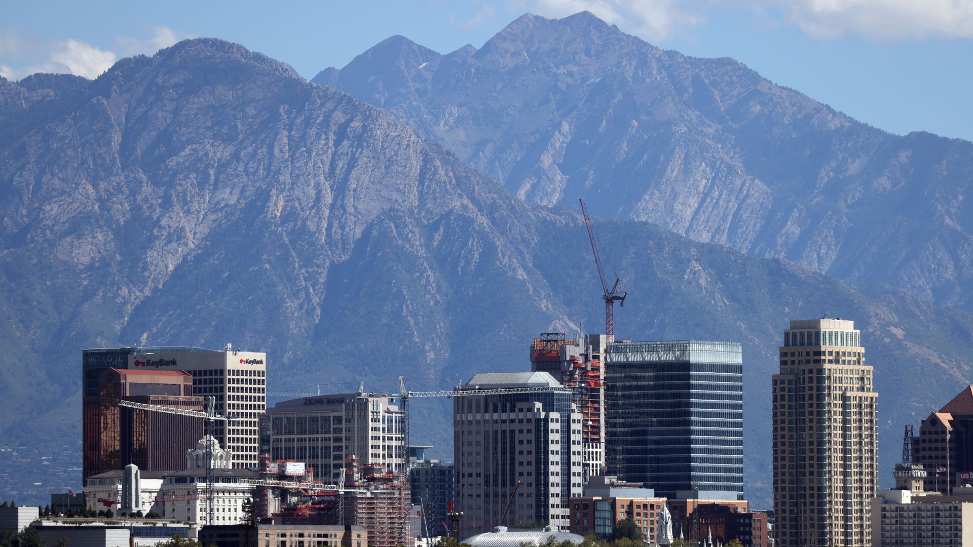 A city skyline with tall glass office towers and a construction crane in the foreground, mountains loom in the distance under a clear blue sky.