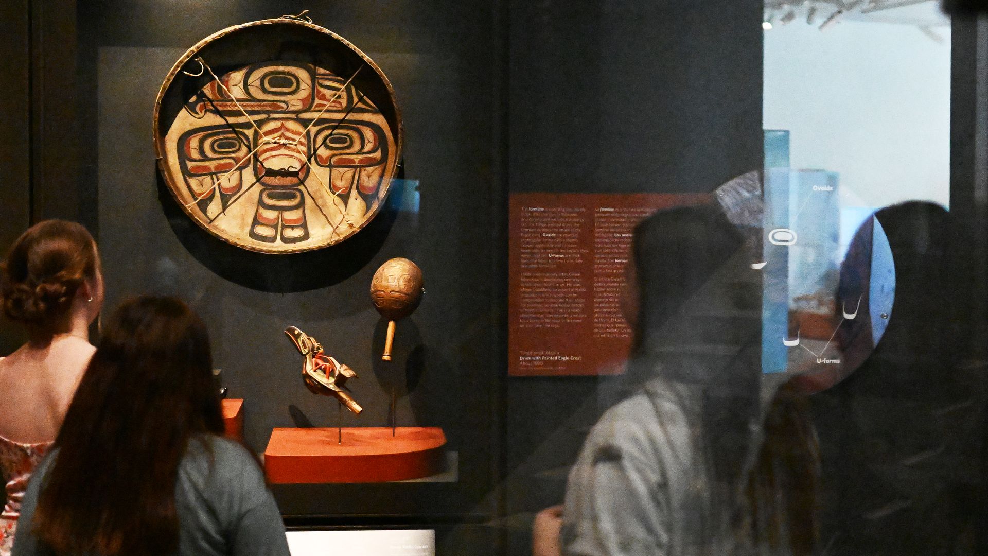 A piece of Native American art hands behind a pexiglass wall while people look on.