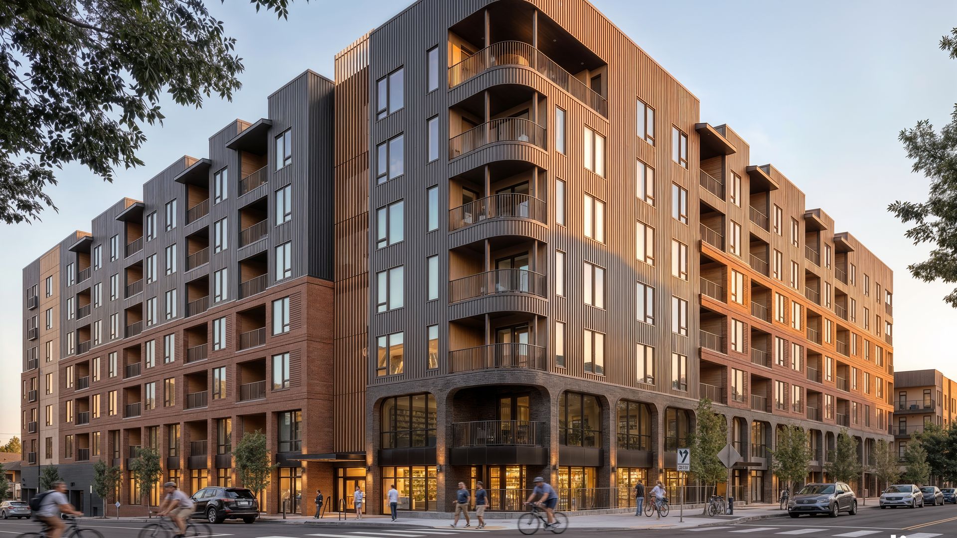 Modern, multi-story building with dark vertical metal siding, curved balconies, and brick base on a busy street. Cyclists and pedestrians pass by as trees frame the scene against a clear blue sky.