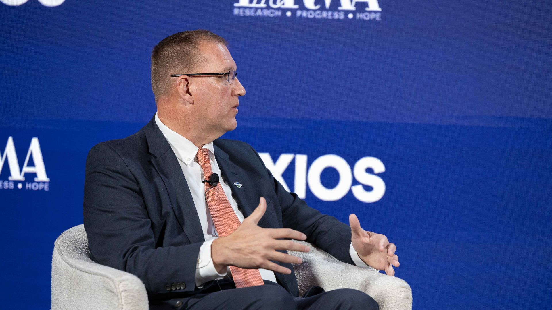 Side profile of a man in a dark suit and pale orange tie, wearing glasses, seated on a stage with a blue backdrop and white text; he gestures with his hands during a discussion.