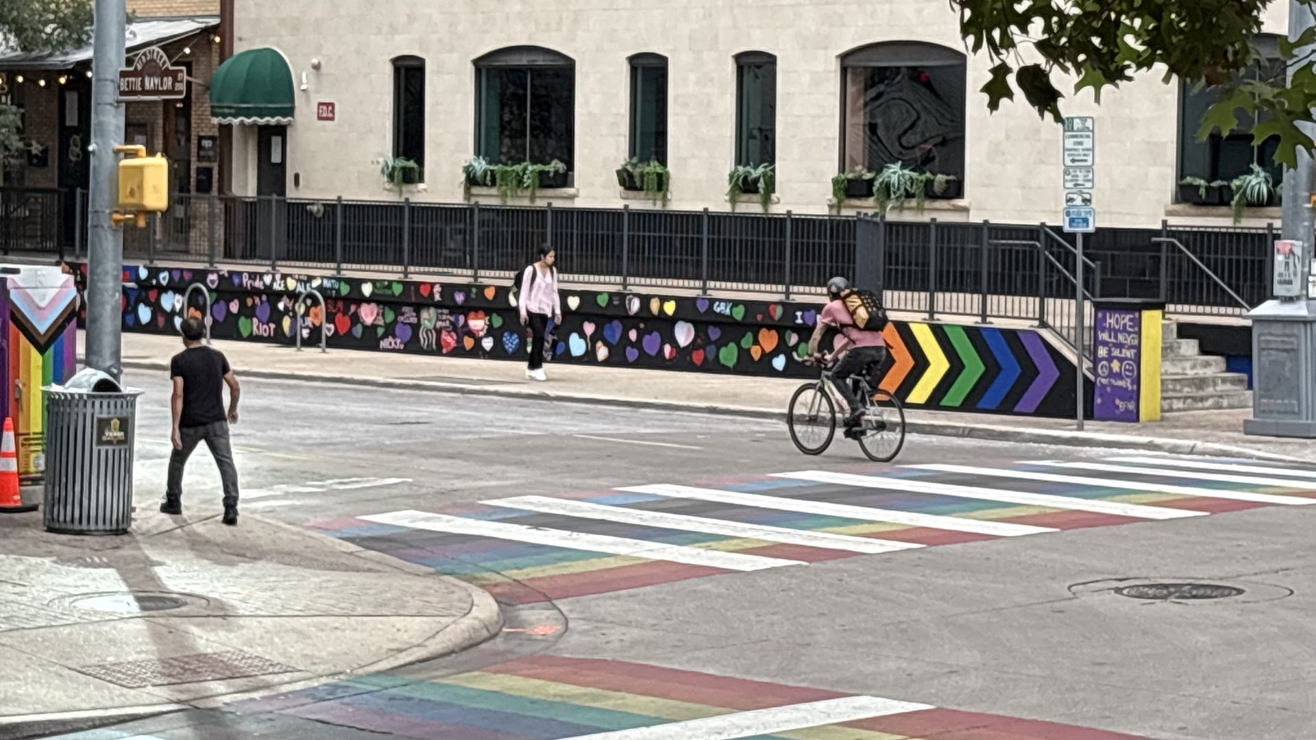 City street scene with a vivid rainbow crosswalk and a wall of colorful heart murals. A cyclist rides by while a pedestrian walks, beside a beige brick building with trees overhead and storefronts in the background.