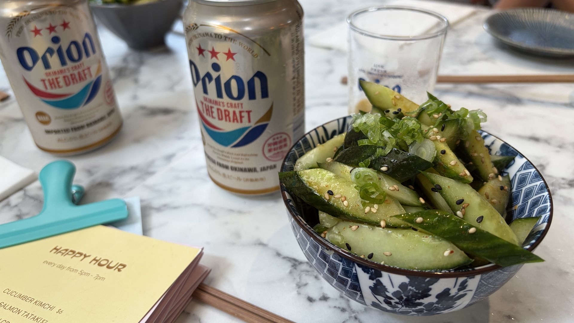 Two Orion beer cans sit on a marble table beside a cucumber salad in a blue-white patterned bowl, with chopsticks, a glass, and yellow menus blurred in the background.