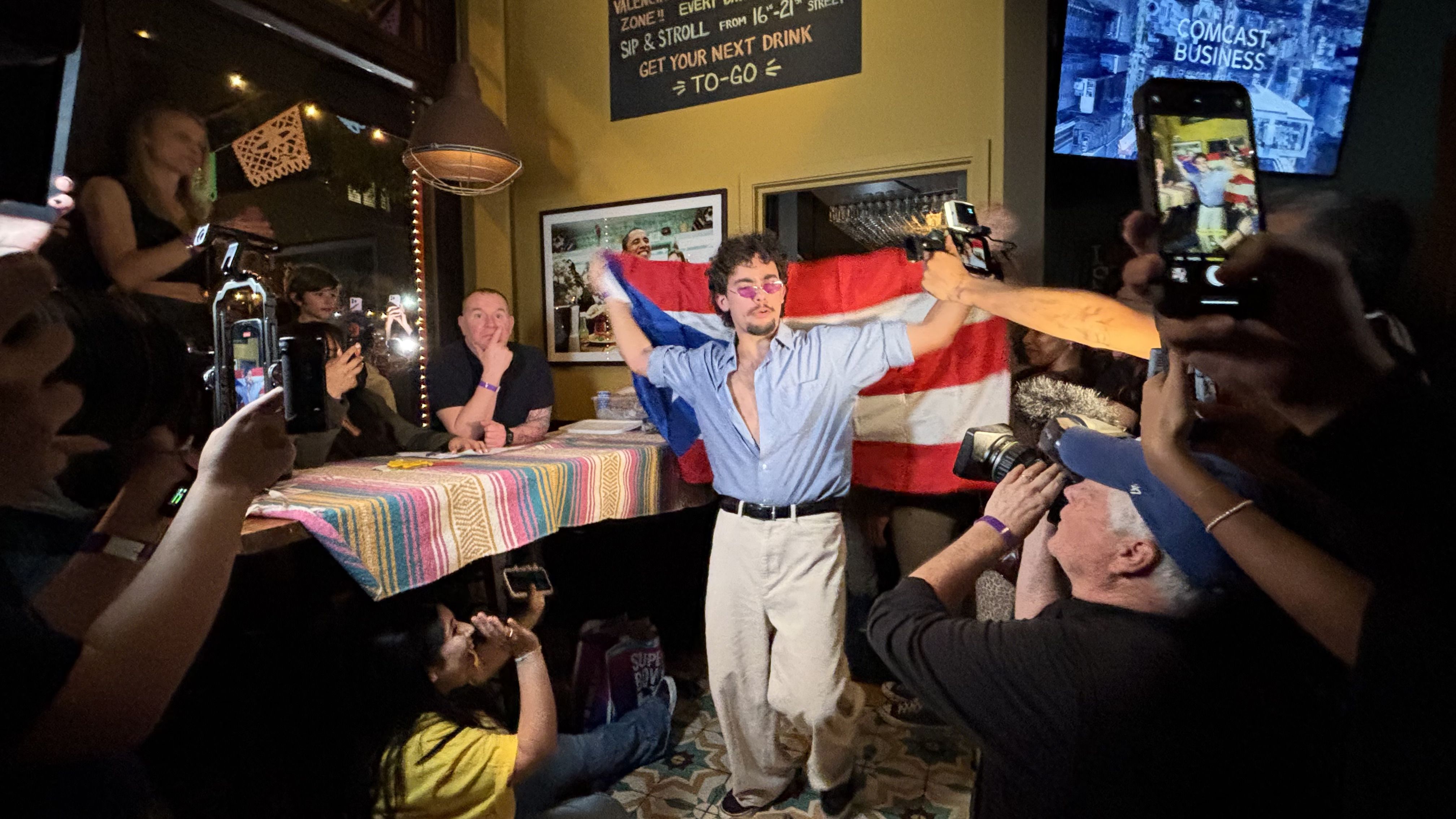 Man with curly hair and purple sunglasses draped in a red, white, and blue flag, posing amidst a crowd taking photos inside a dimly lit room with colorful tablecloth and wall signs.