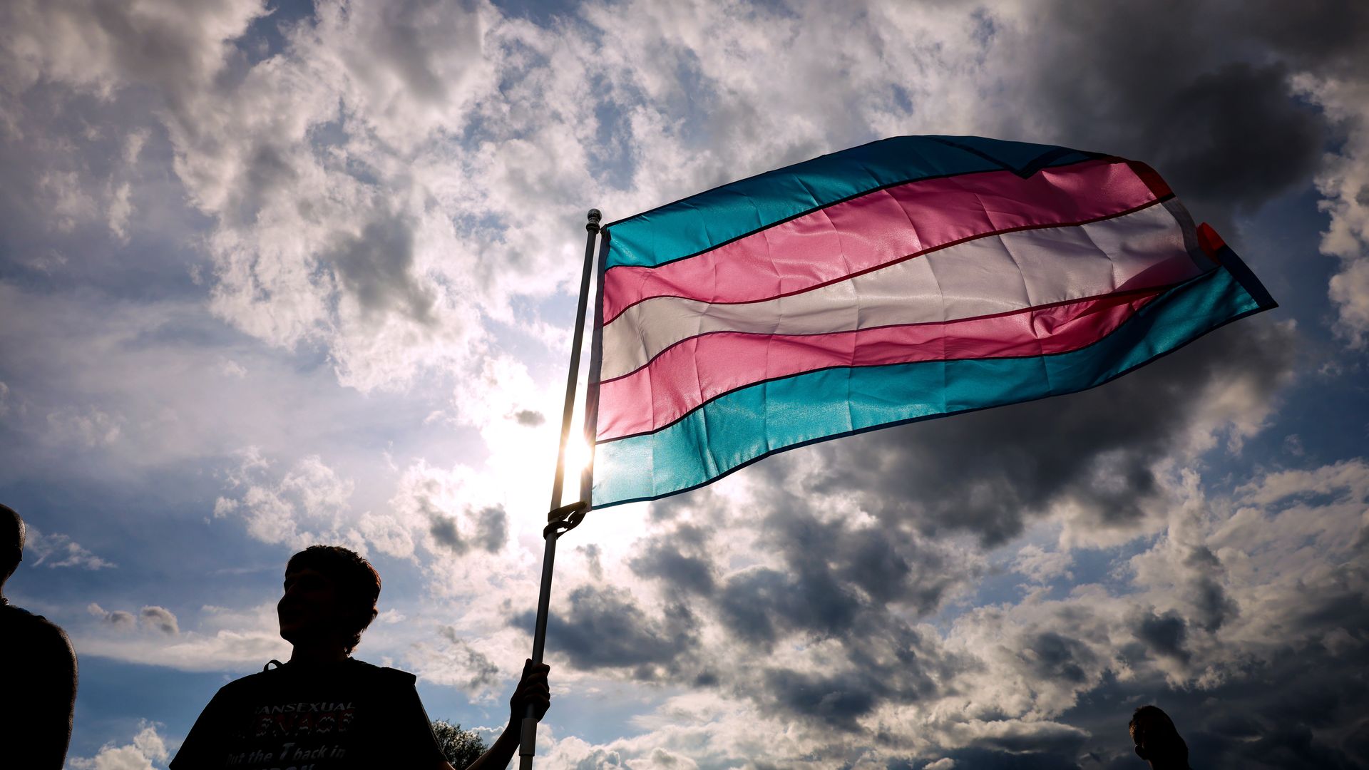 A person holds a transgender flag (blue, pink, white) with a cloudy sky in the background
