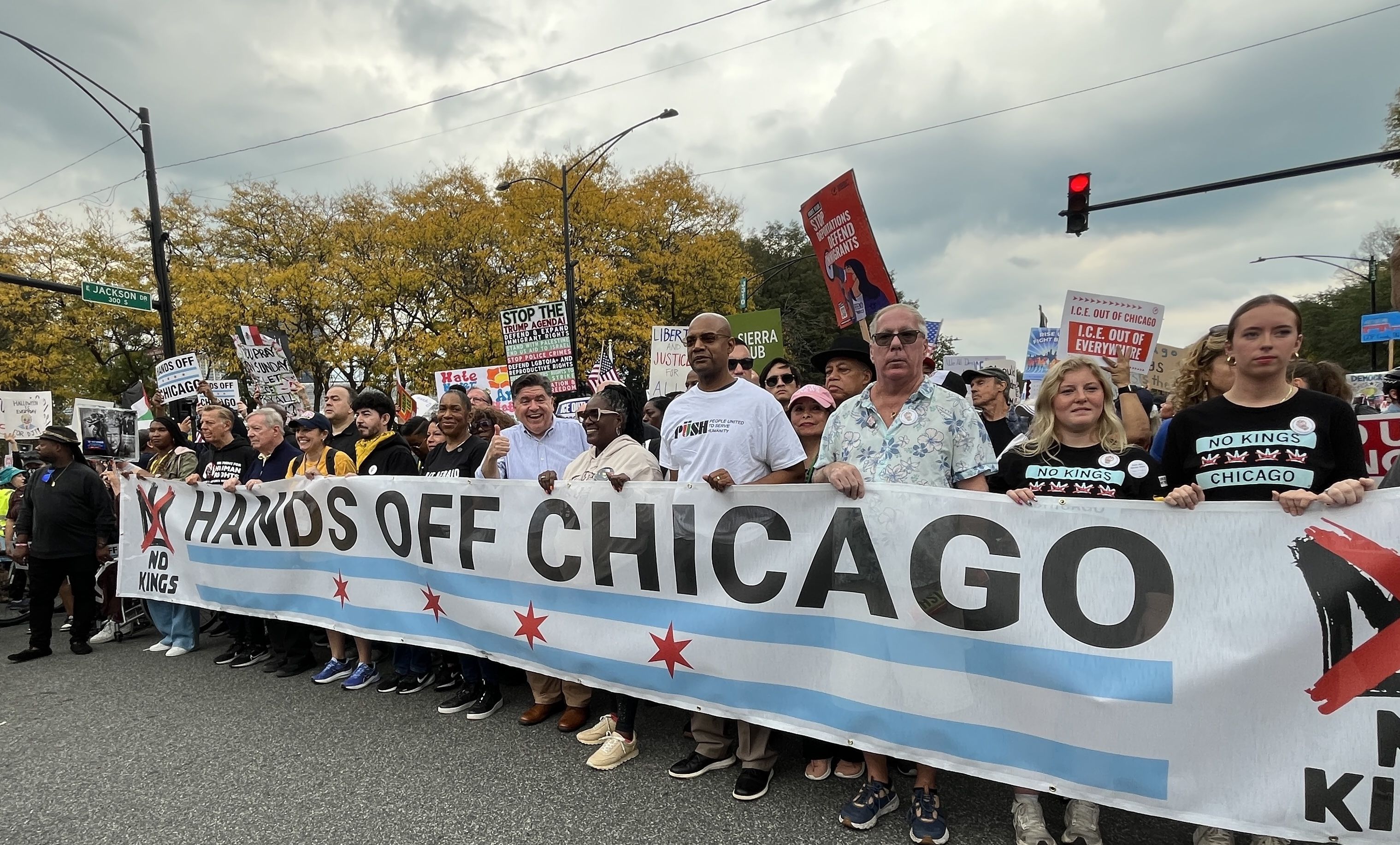 Group of protesters including Illinois Gov. JB Pritzker holding a large banner reading "HANDS OFF CHICAGO" with signs about immigration and no kings, under cloudy sky and autumn trees on city street.