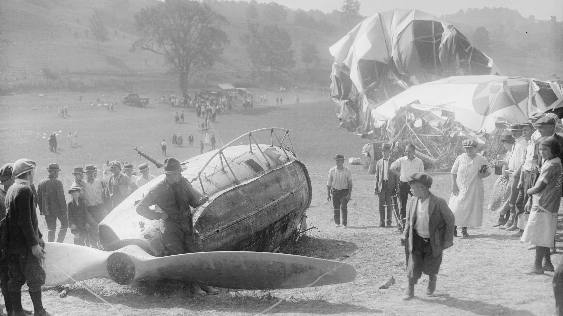 Historical black and white photos of people in a field gathered around the wreckage of a downed rigid airship, with a propeller, metallic frame and exterior tarpaulin visible.