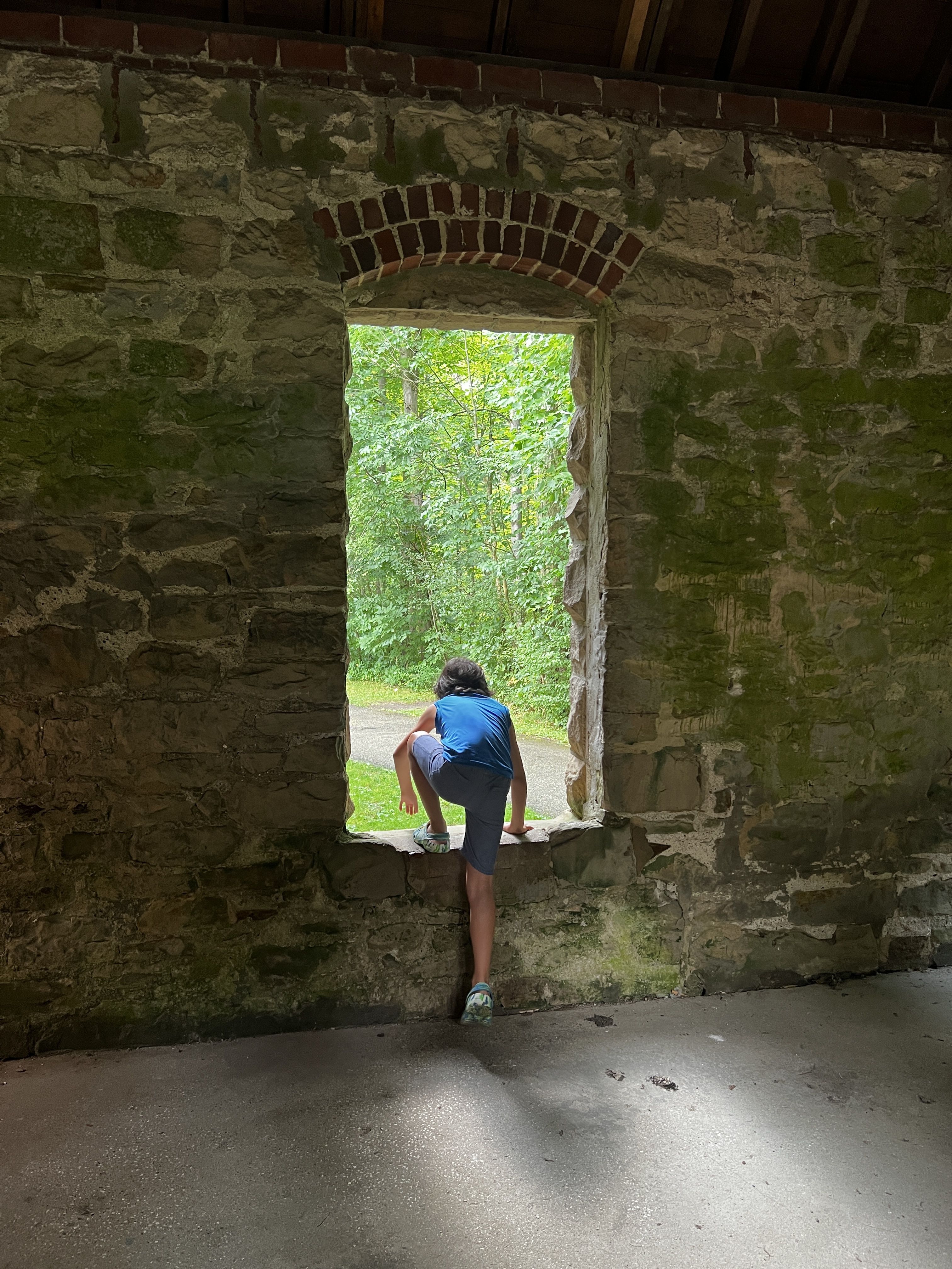 Inside a mossy stone ruin, a person in a blue shirt crouches on a rough windowsill, peering through a brick-arched opening at bright green trees outside.