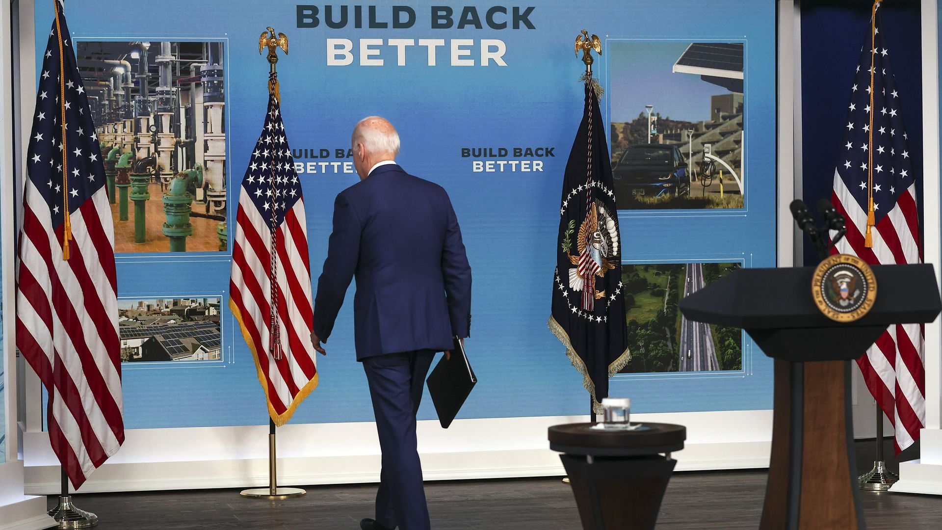 A view of President Biden from behind walking away from a podium with a "Build Back Better" backdrop