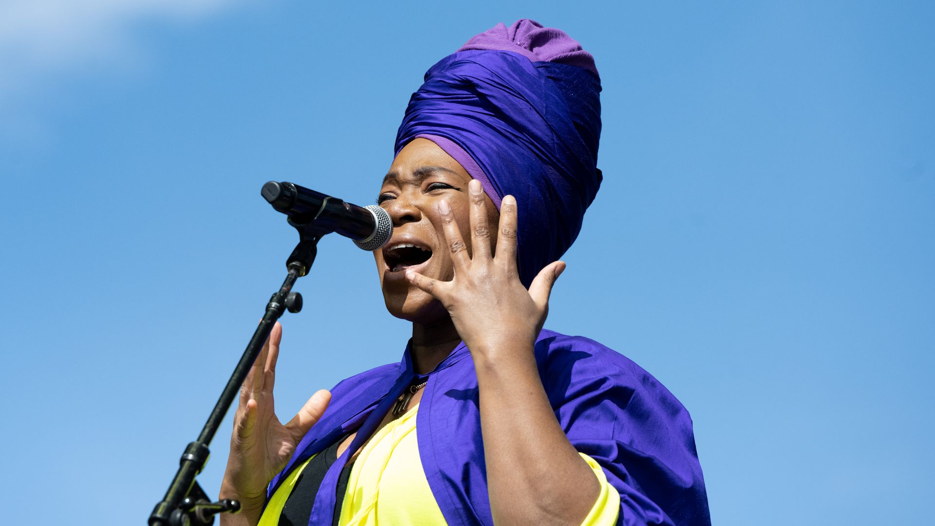 India.Arie wears a bright purple and yellow wrap while singing against a blue sky background