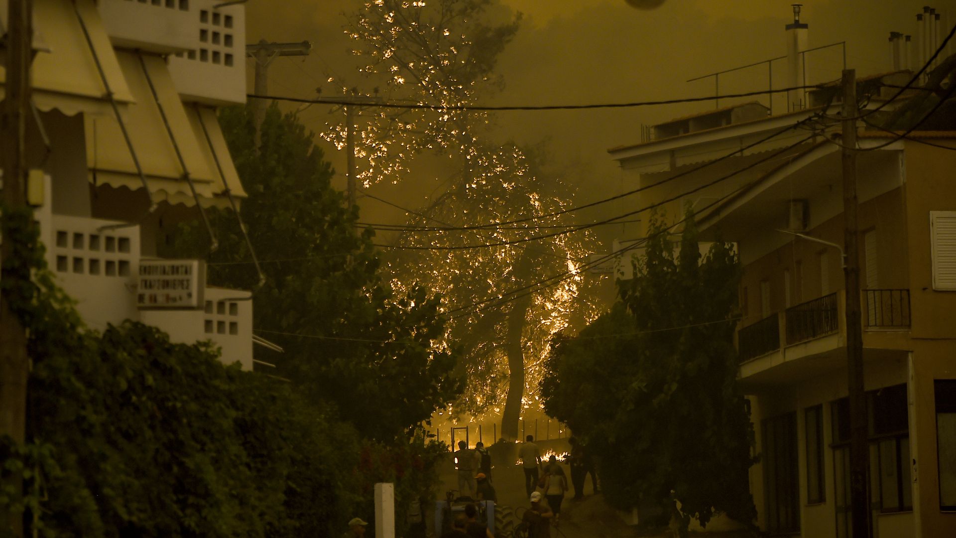 Photo of a fire burning in the distance as a crowd watches from the street of a town