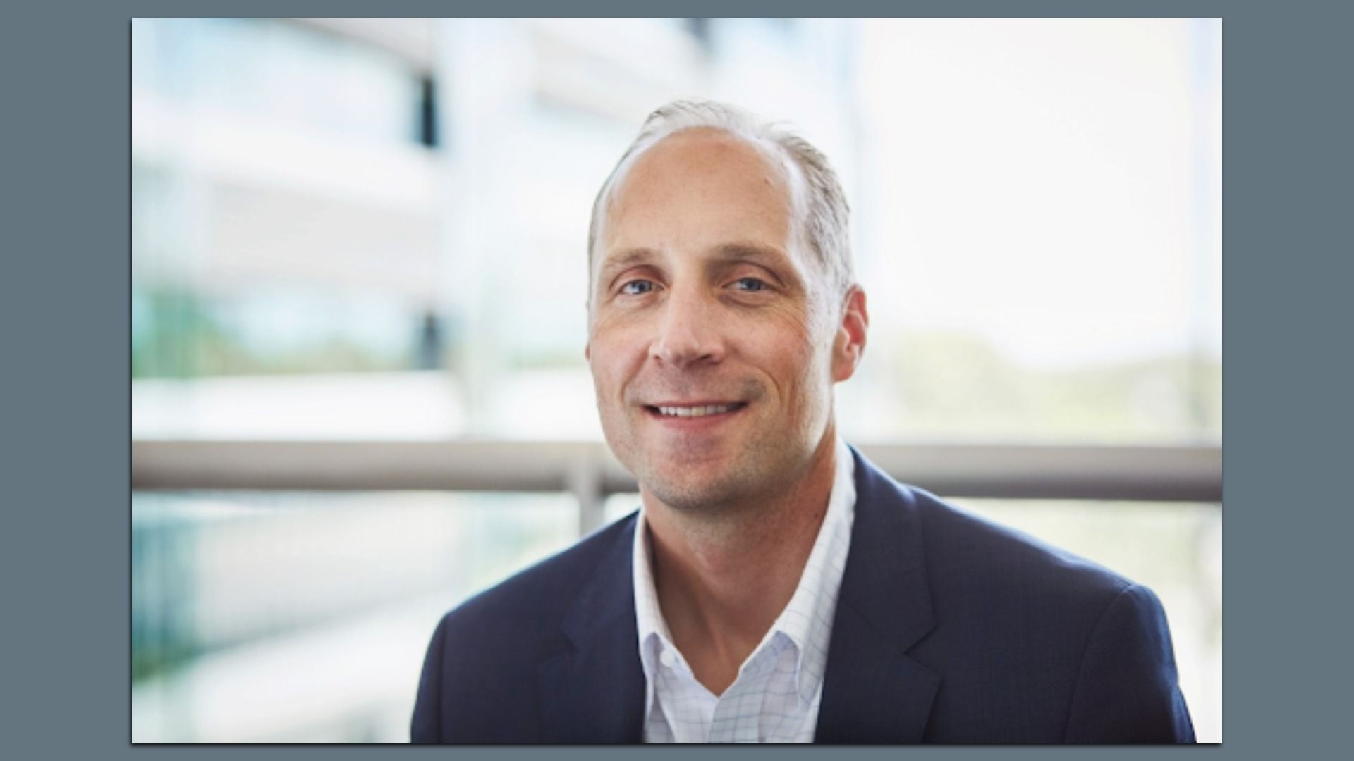 Portrait of Chad Bown, a smiling middle-aged man with gray hair wearing a dark blazer and white shirt, posed in a bright, modern office with glass windows in the blurred background.