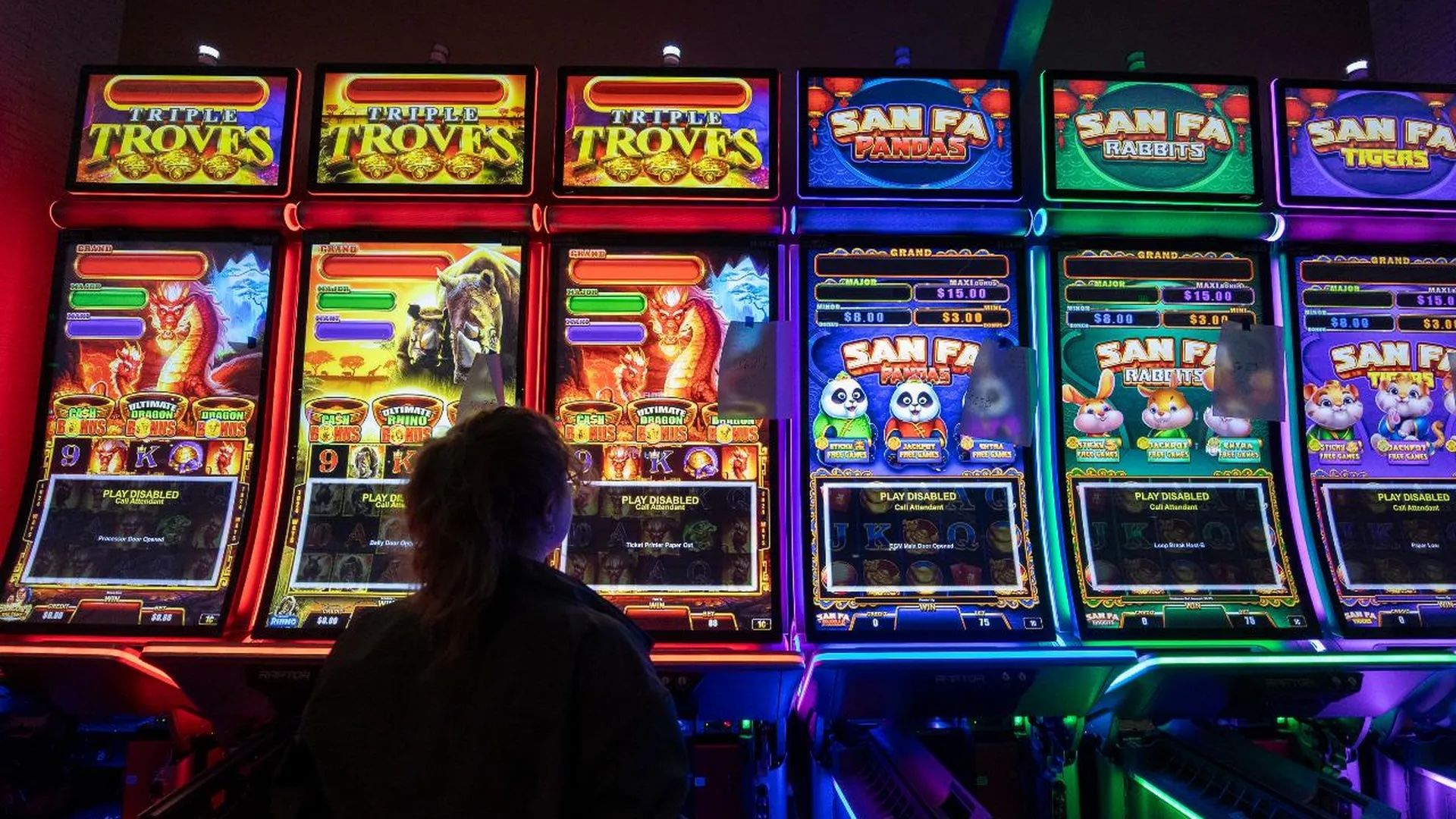 Row of neon-lit casino slot machines with bright, colorful screens showing Triple Troves and other game titles, a person stands with back to camera in front of them under red, blue and green lighting.