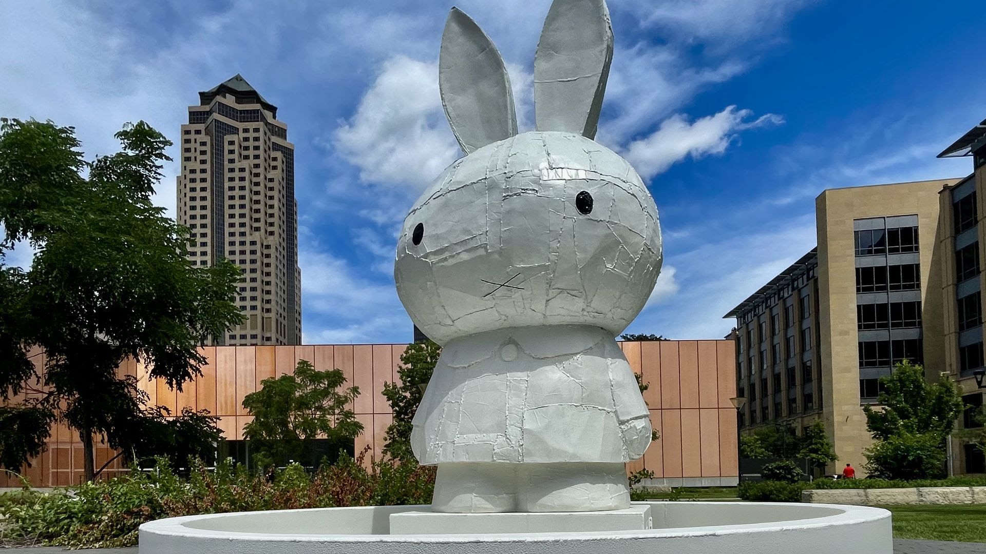 Large white patchwork bunny sculpture with tall ears, stitched seams, and black eyes, perched on a pedestal in a circular fountain basin, against blue sky, greenery, and city buildings.