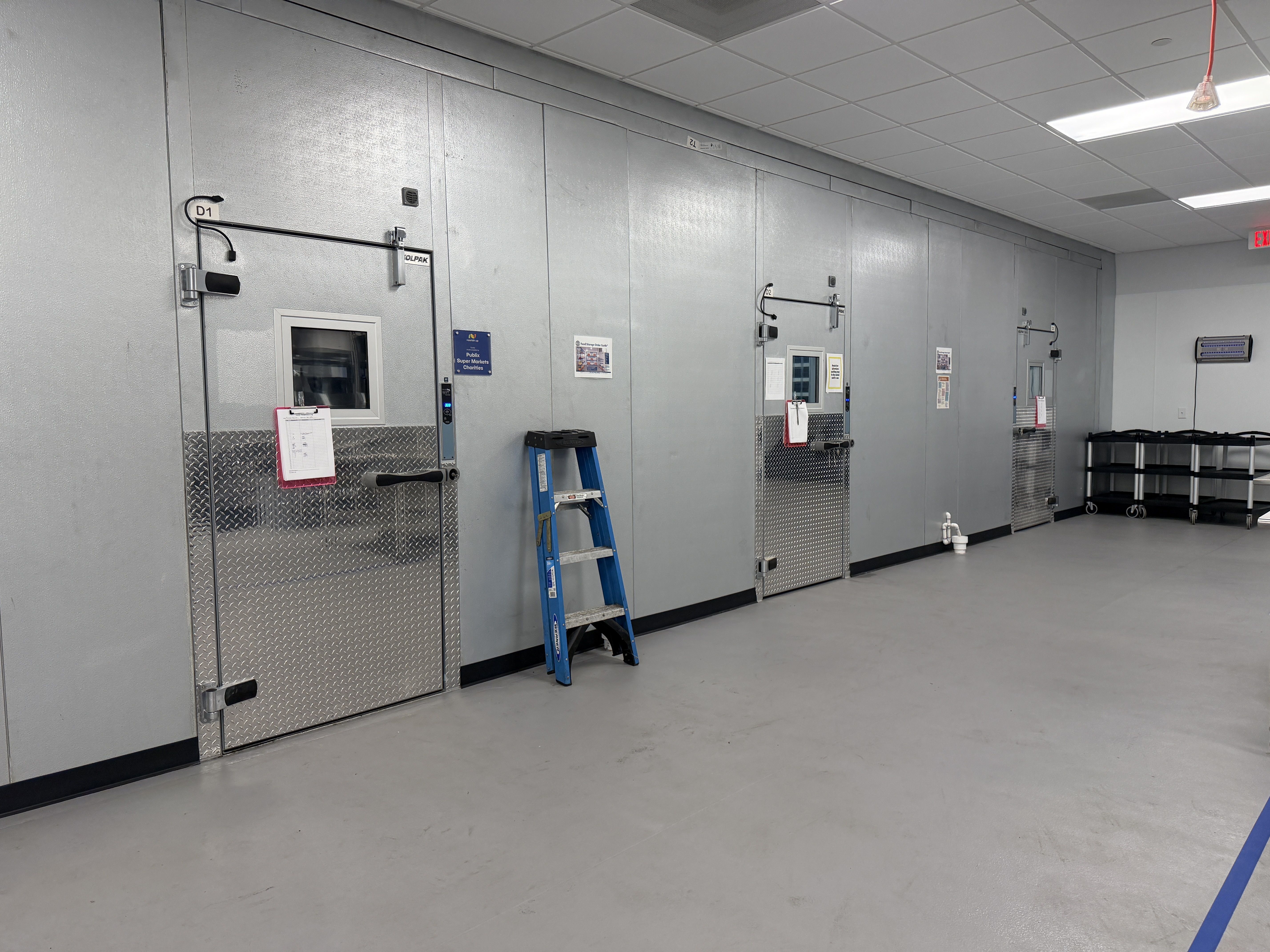 Industrial cold storage facility with tall gray insulated doors along a wall, labeled D1 and D2. Diamond metal panels at the base, a blue ladder, clipboards, and overhead fluorescent lights.
